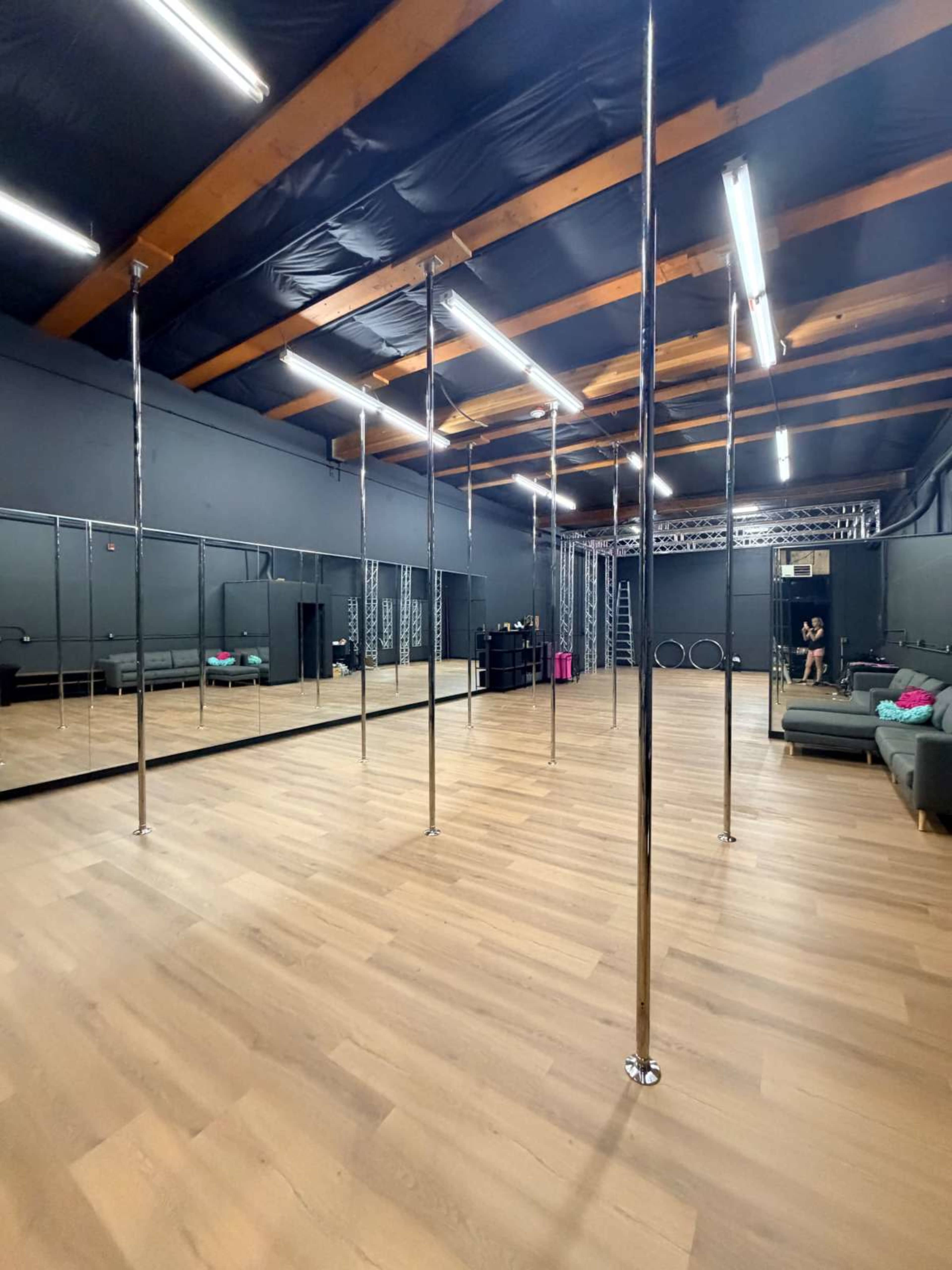 The image shows a spacious studio with polished wooden floors, mirrored walls, and several vertical poles, all under a black ceiling with exposed wooden beams.