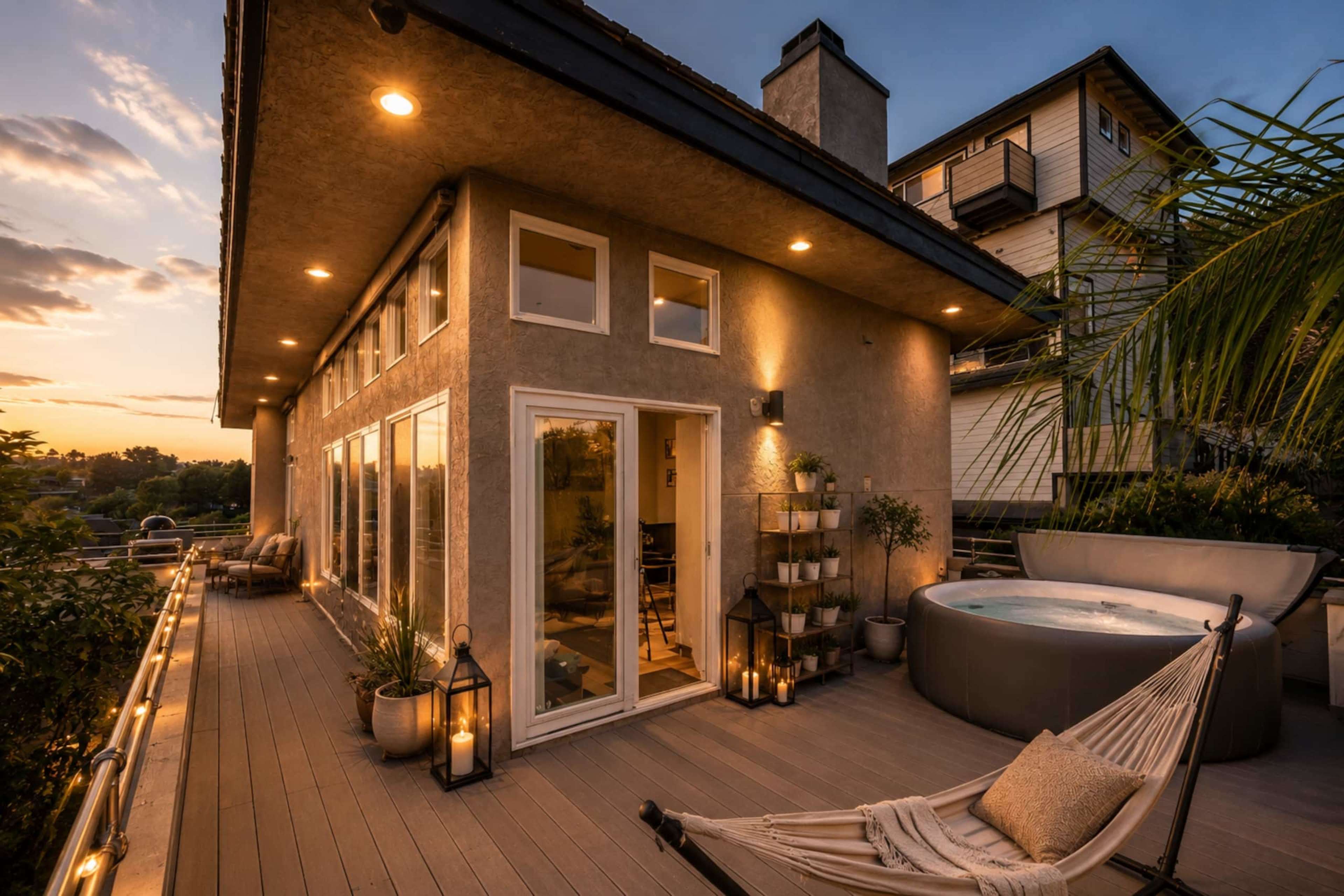A modern home features large windows, a deck with a hot tub, and potted plants, all illuminated by outdoor lighting during sunset.