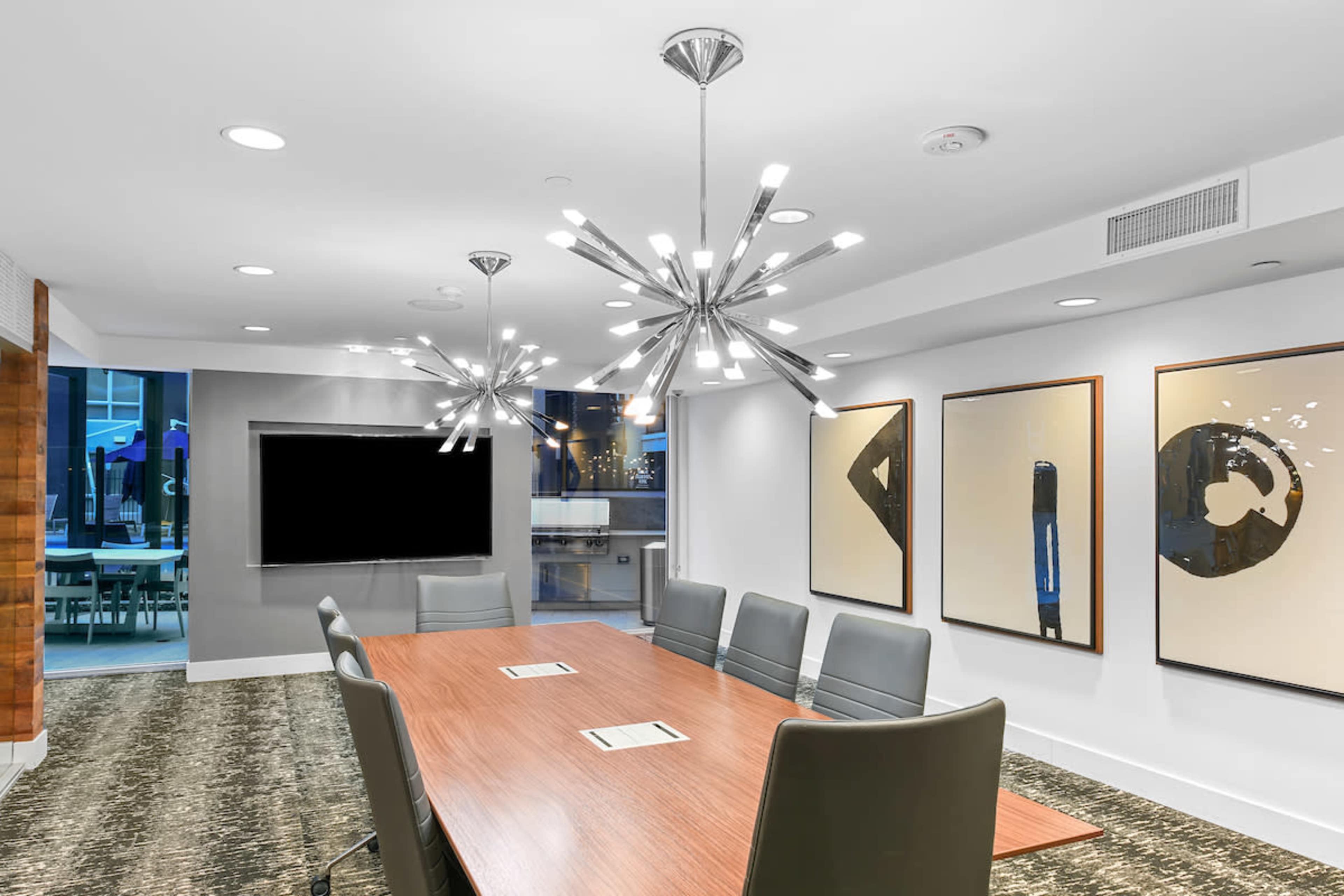 A modern conference room features a long wooden table surrounded by ergonomic chairs, with two contemporary light fixtures and large abstract artworks on the walls.