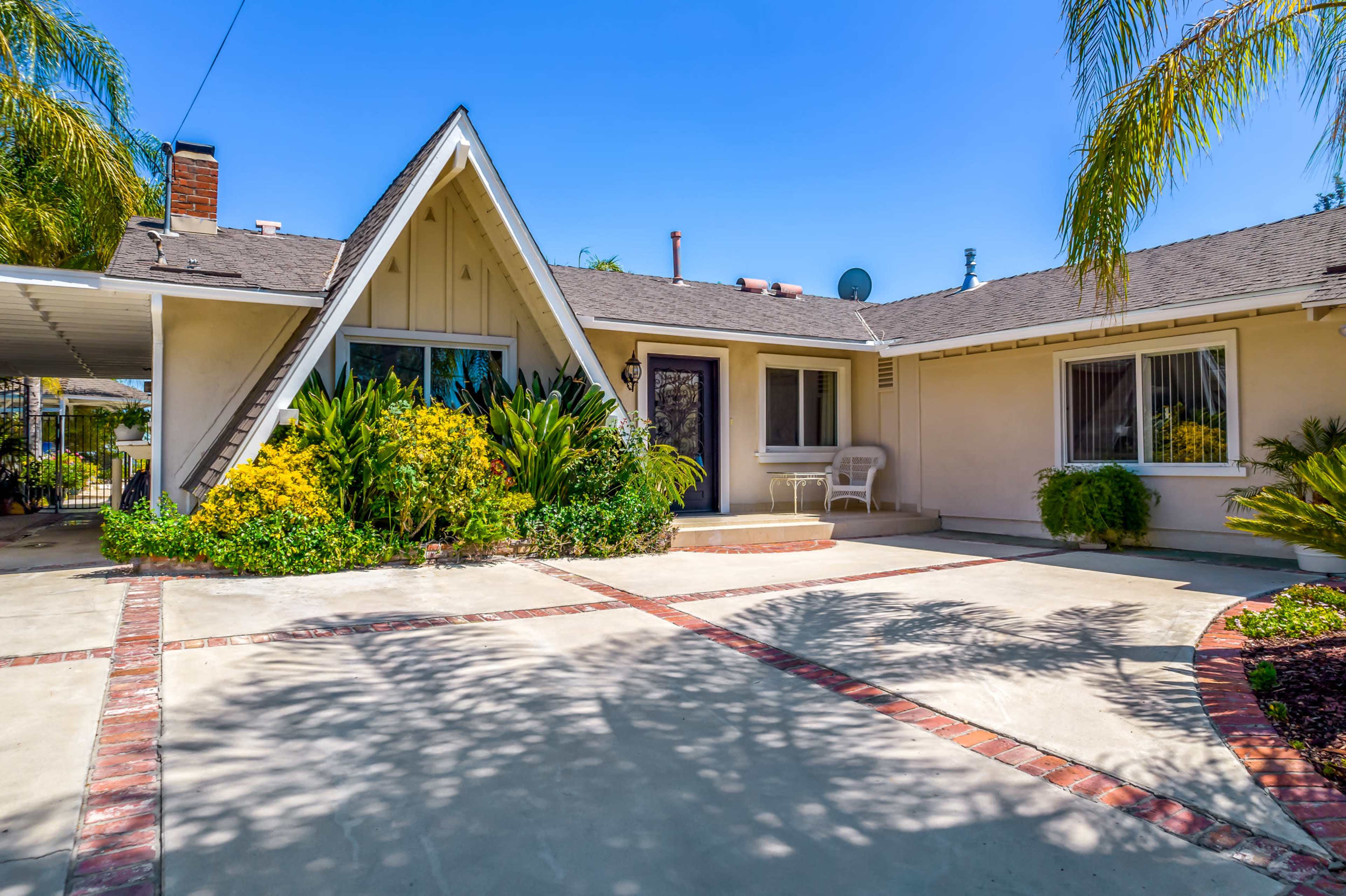 A single-story A-frame house with a concrete driveway and a landscaped front yard featuring various plants.