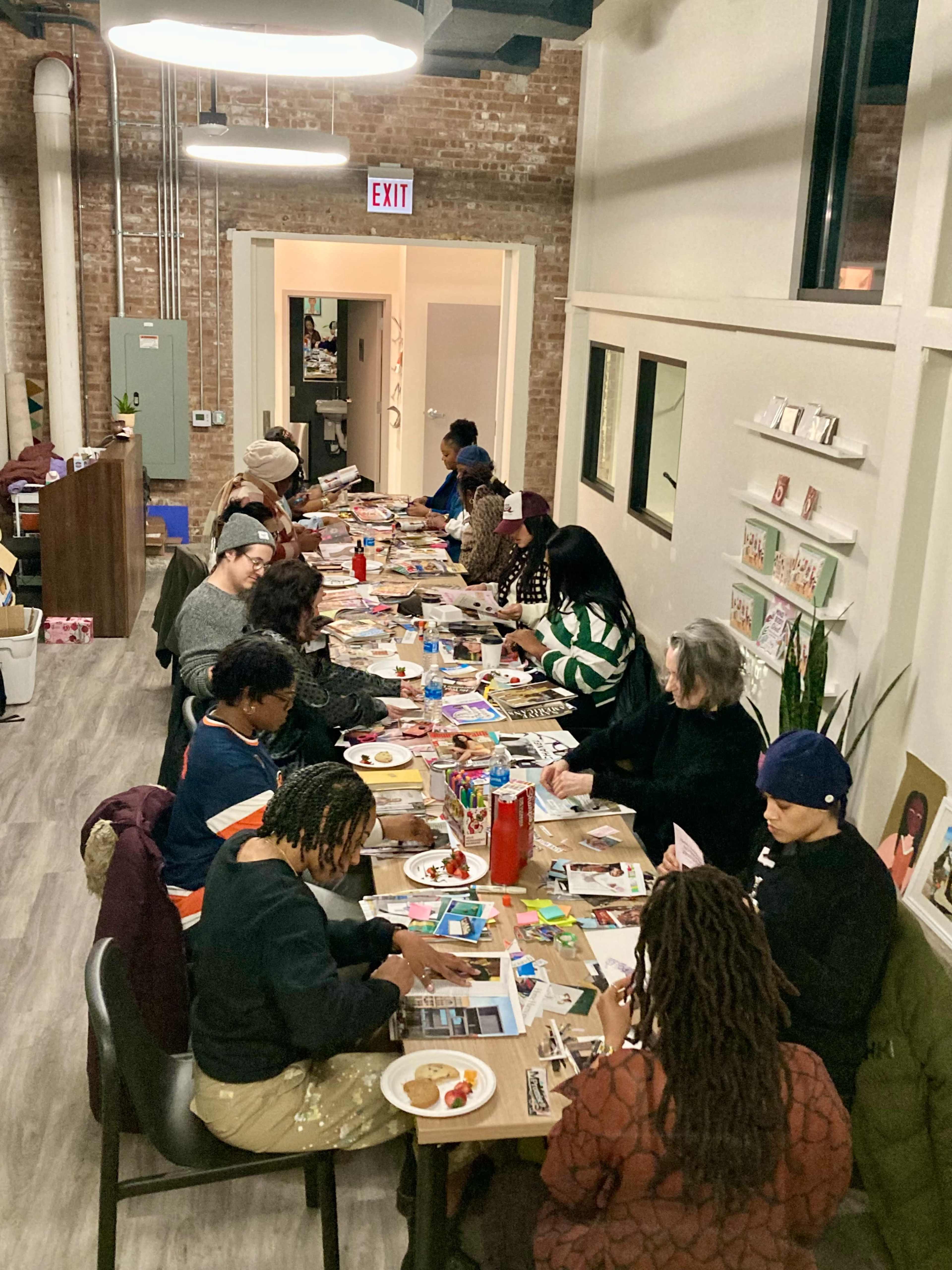 A group of people is seated at a long table, engaged in creative activities with various materials spread out in a room featuring exposed brick walls and large windows.