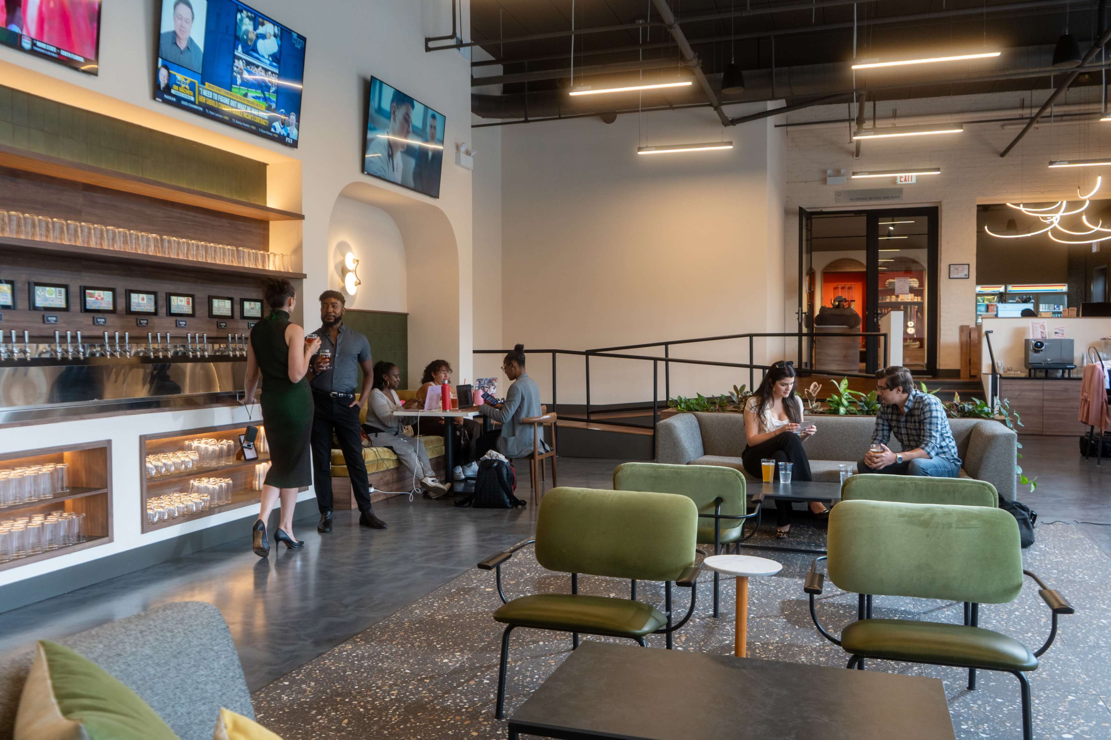 The image shows a modern workspace with a well-lit lounge area featuring seating, a beverage station, and several groups of people engaged in conversation or work.