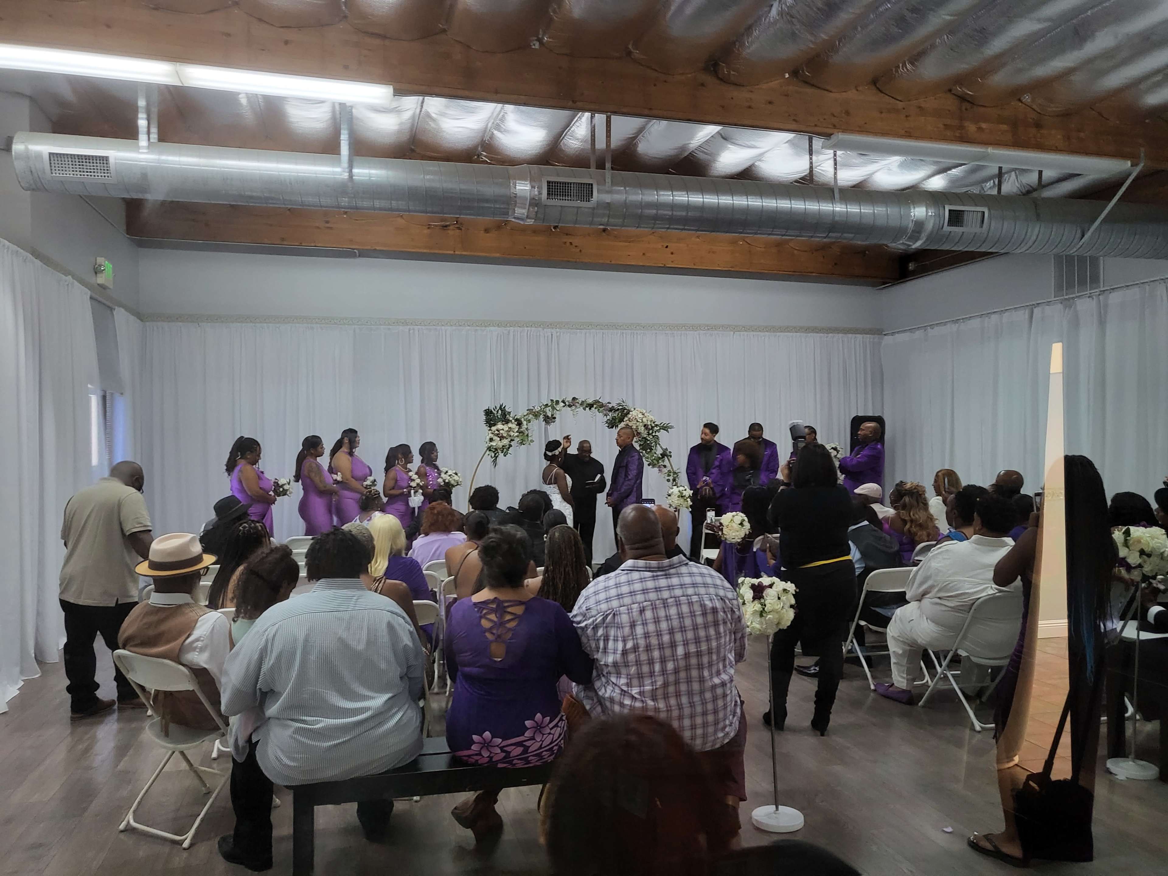 A wedding ceremony is taking place indoors with guests seated in folding chairs and a wedding party in purple attire gathered near an arch decorated with flowers.