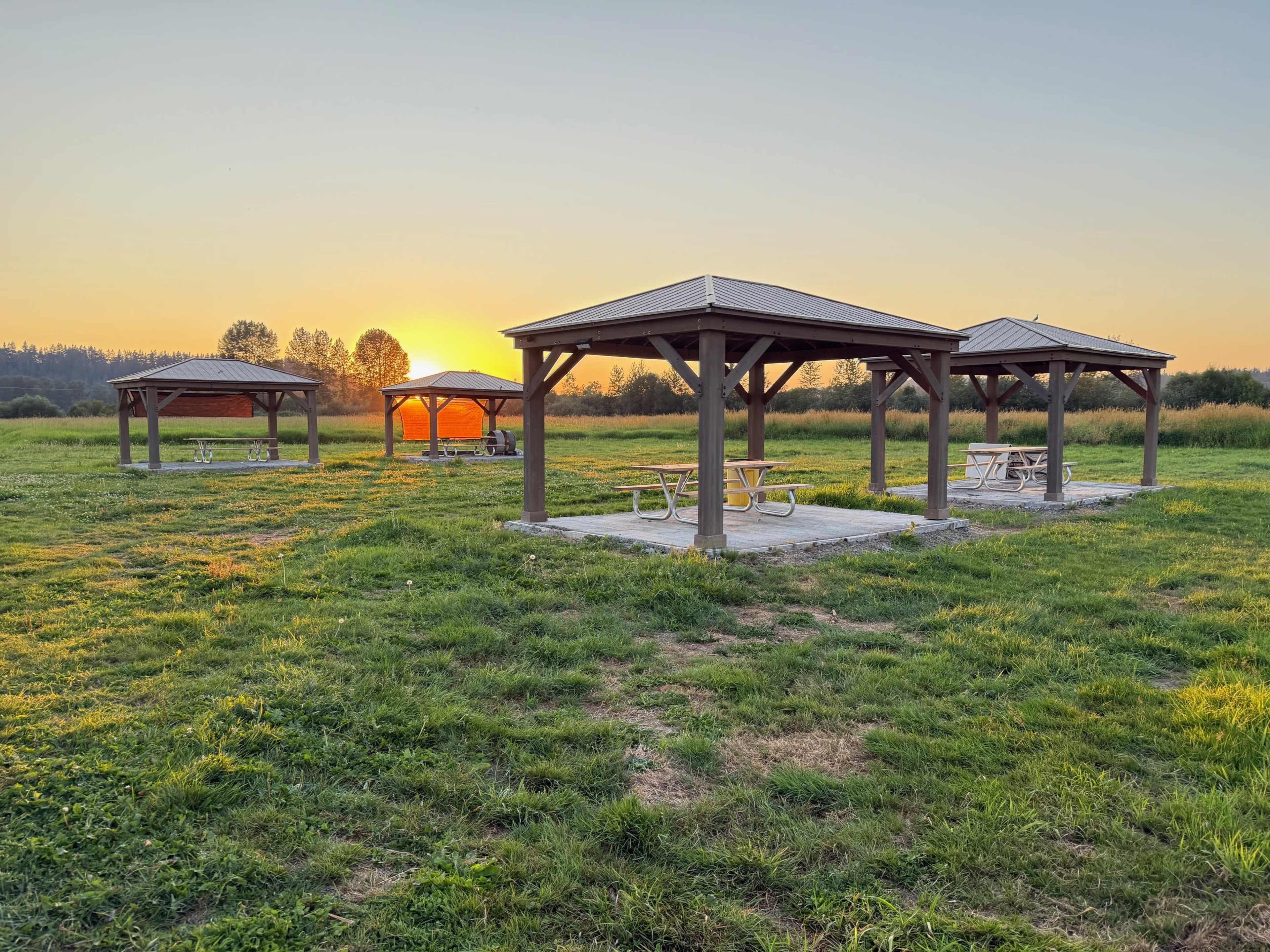 The image shows several wooden pavilions in a grassy area with a sunset in the background.