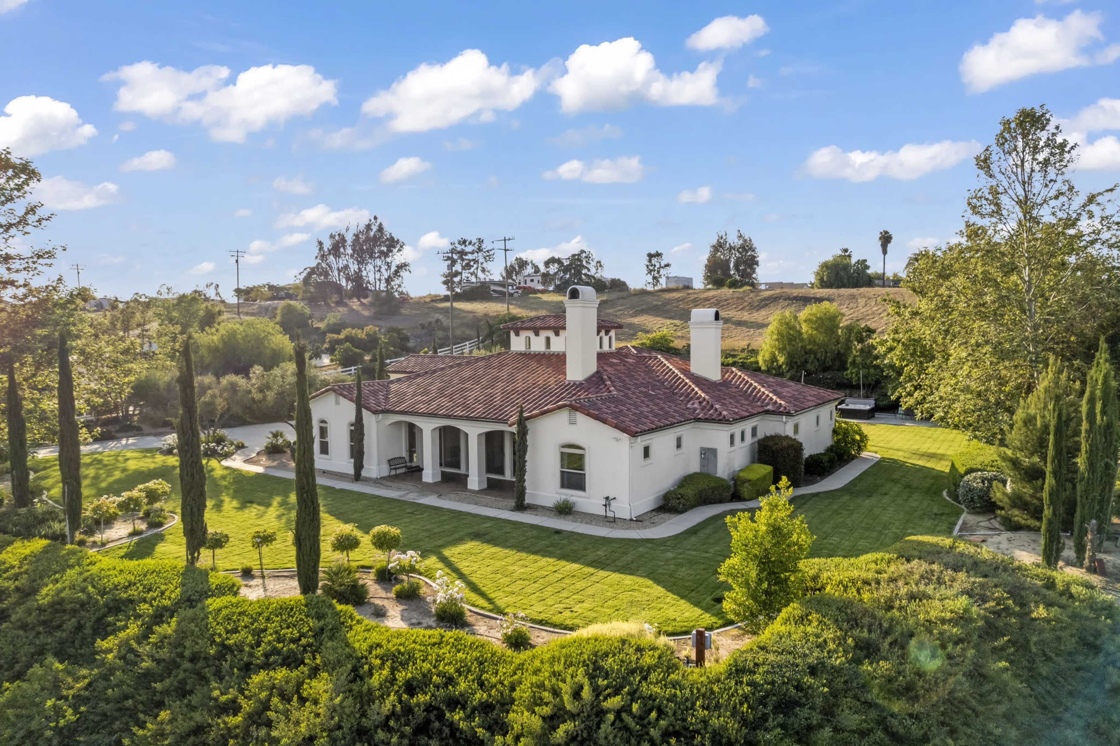 A single-story house with a tiled roof and manicured lawn is surrounded by trees and shrubs in a rural setting.