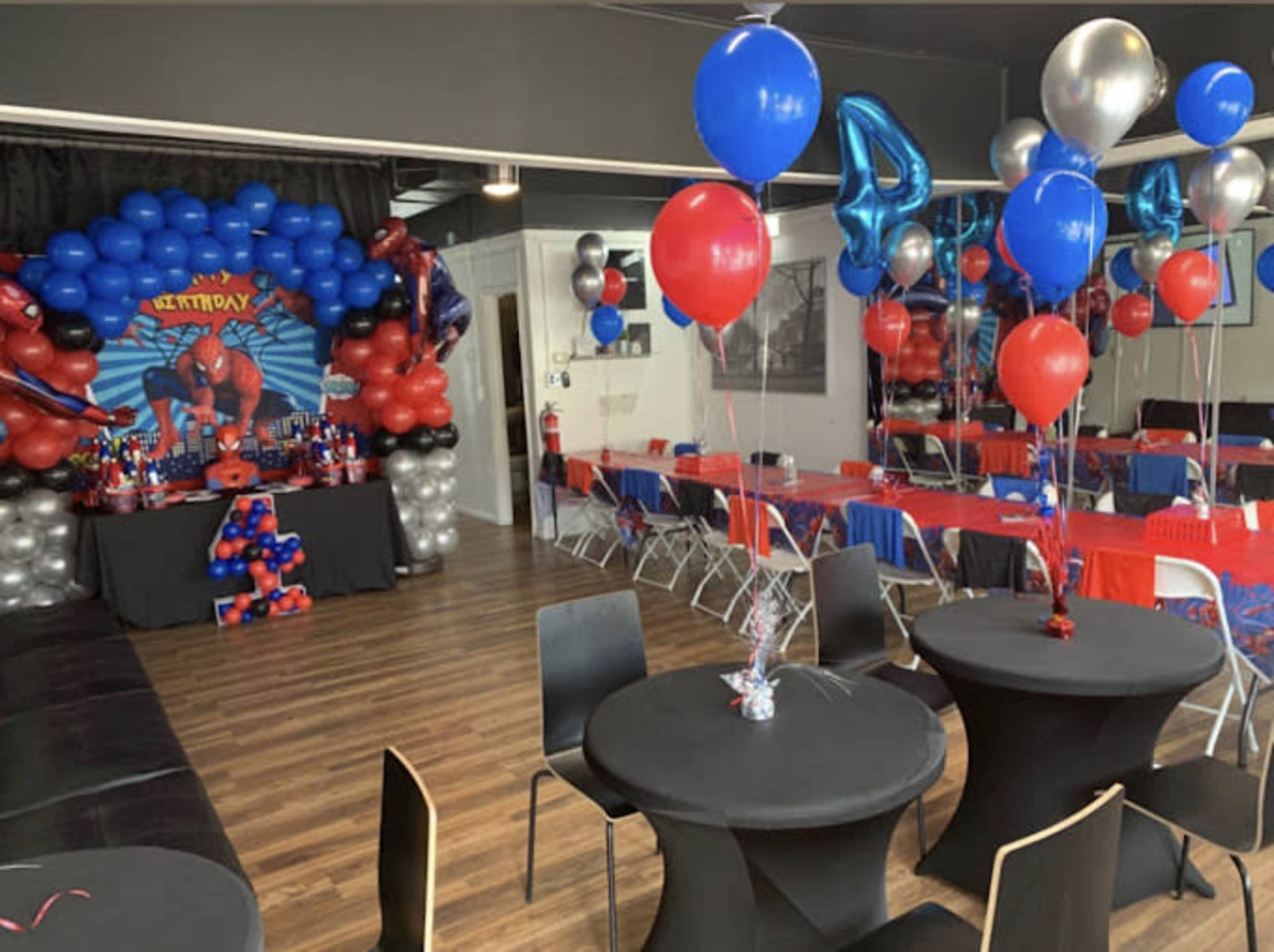 The image shows a decorated party venue featuring red, blue, and black balloon arrangements, tables set with red tablecloths, and a birthday theme centered around a superhero.