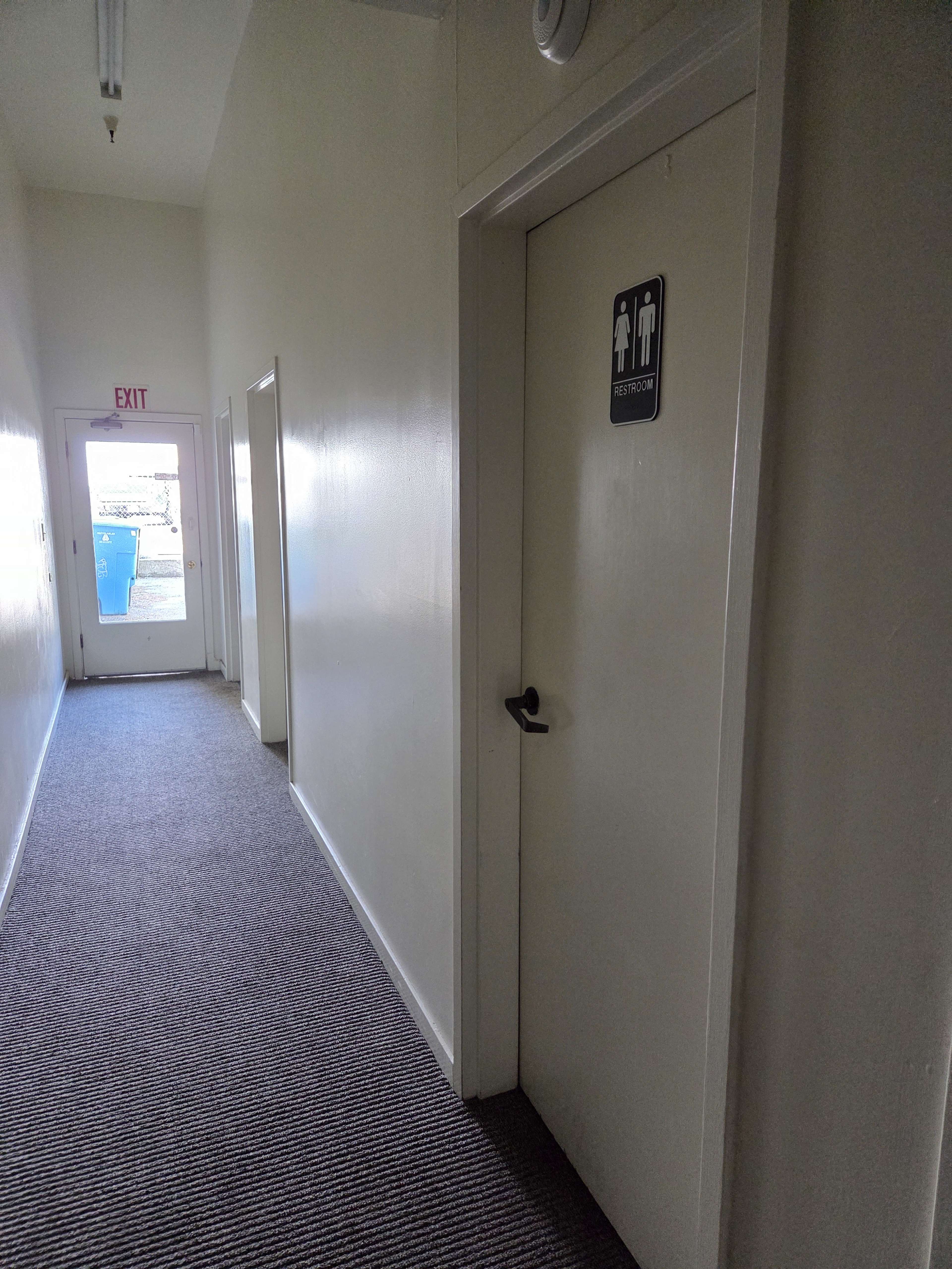 The image shows a narrow hallway with a restroom door on the right side and an exit at the far end.
