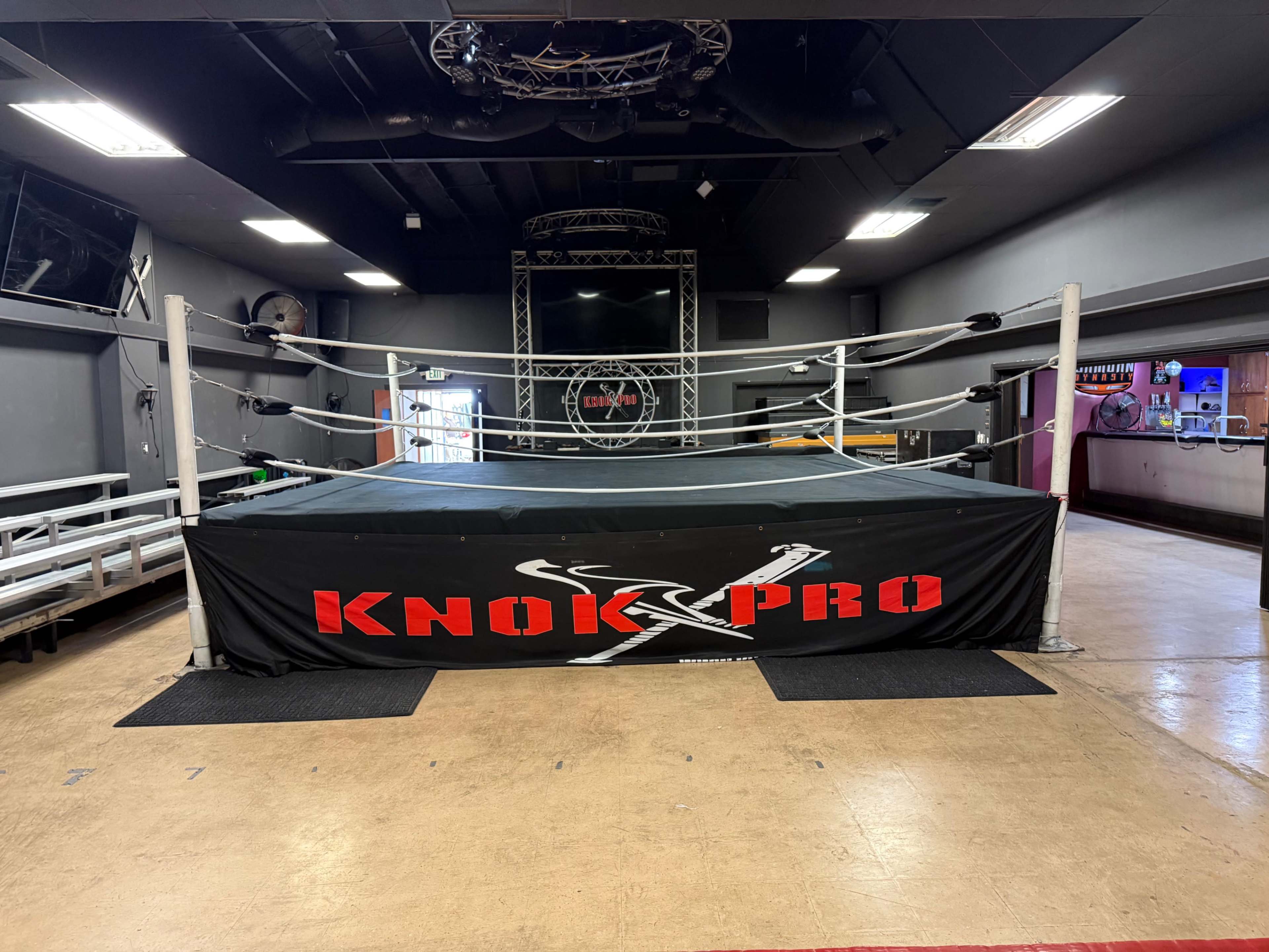 The image shows a professional wrestling ring set up in a training facility, with benches for spectators in the background and a lighting structure above the ring.