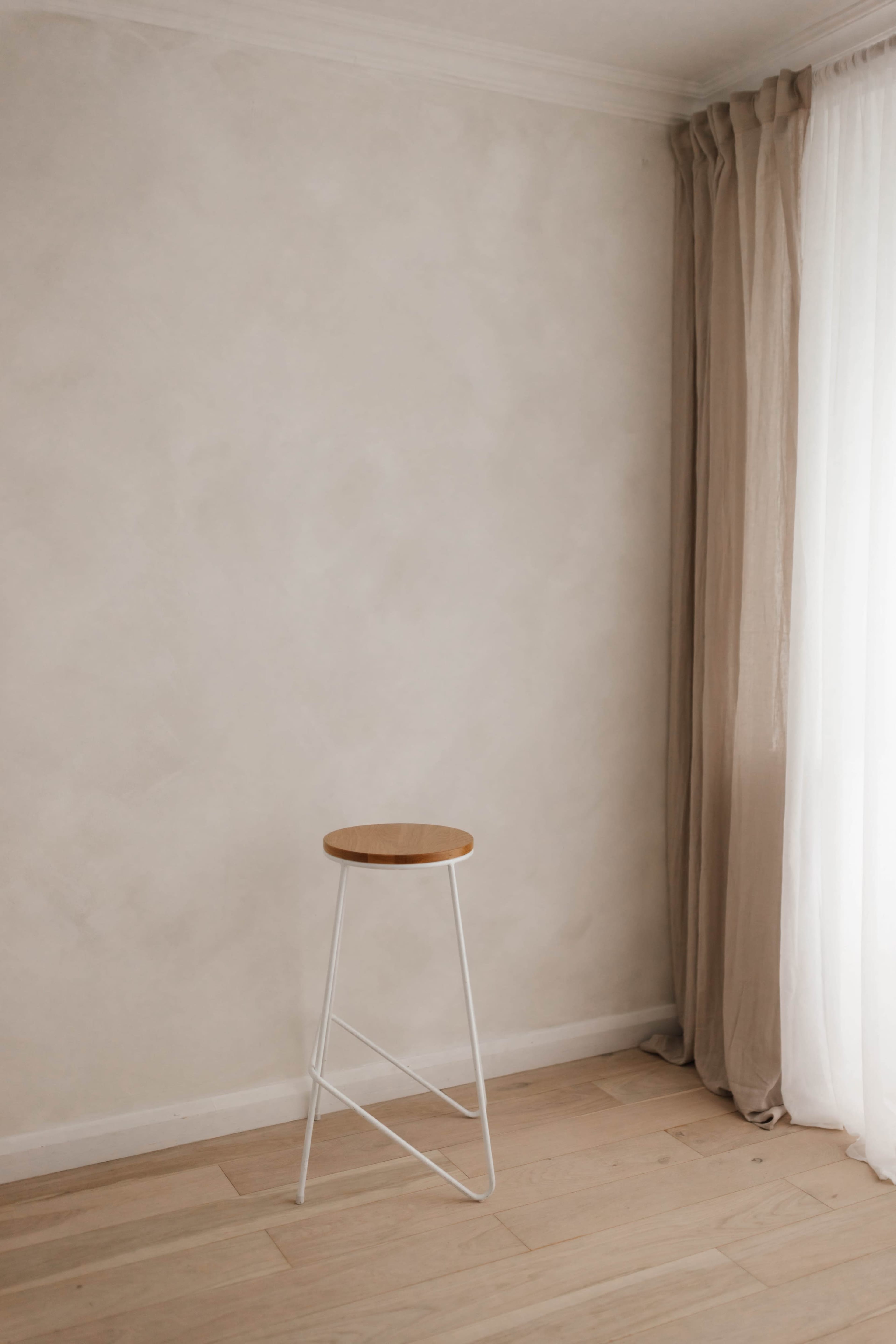 A minimalist space features a single wooden stool against a light-colored wall next to sheer curtains.