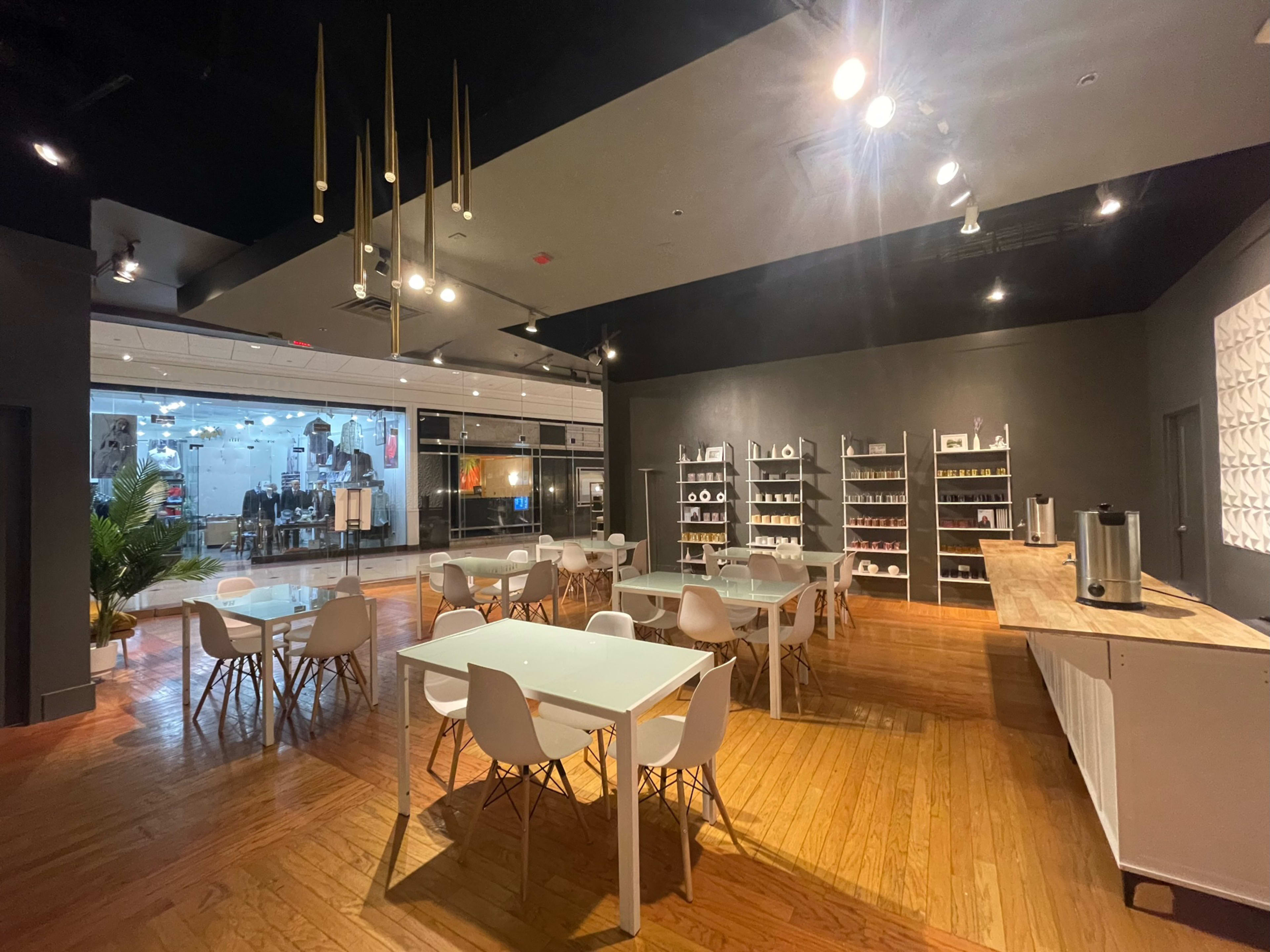 A modern, brightly lit café or meeting space with white tables and chairs, wooden flooring, and a shelving unit displaying dishware against a dark wall.