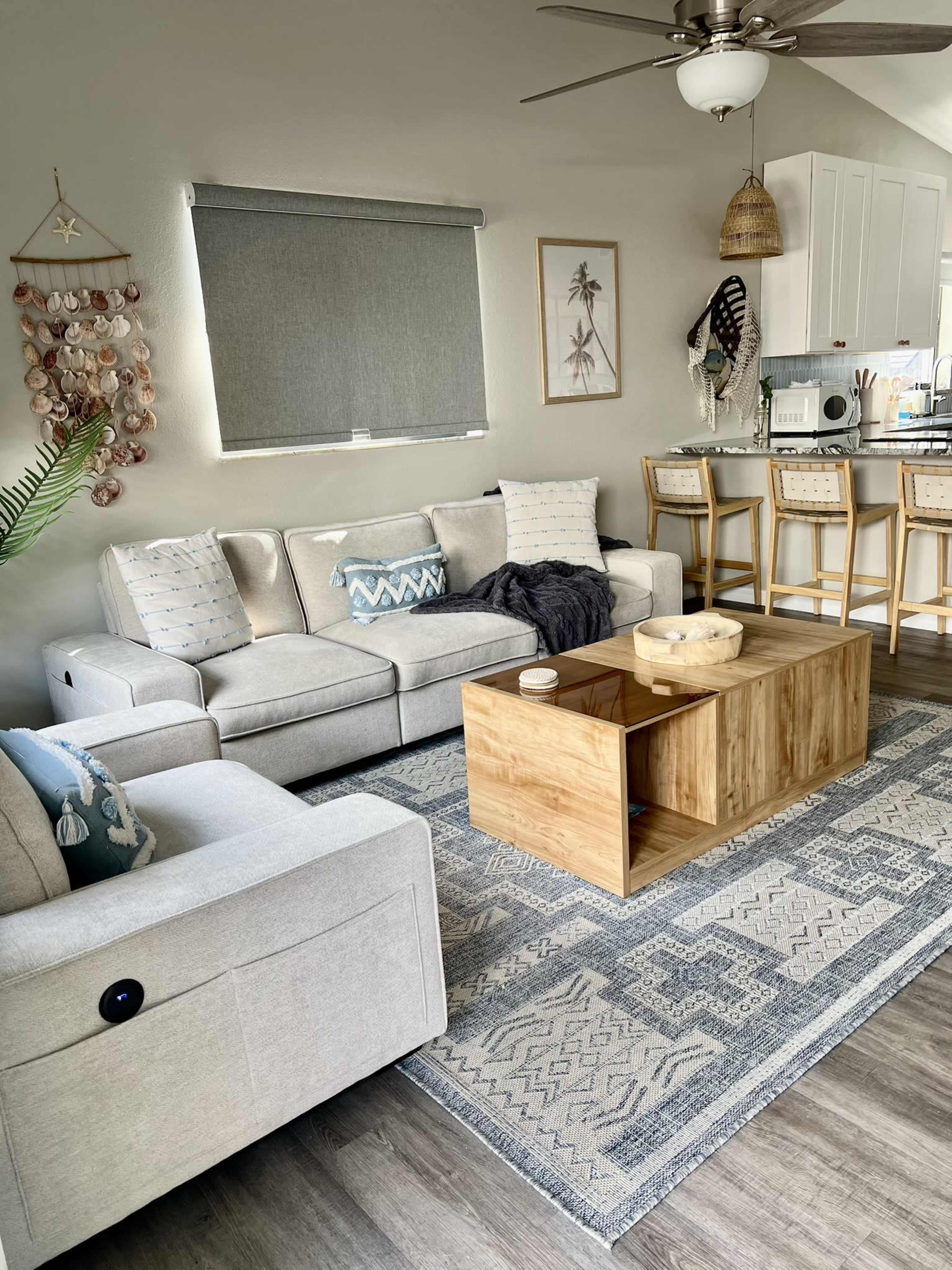 The living room features a light gray sofa and armchair, a wooden coffee table, and a kitchen bar with stools in the background.