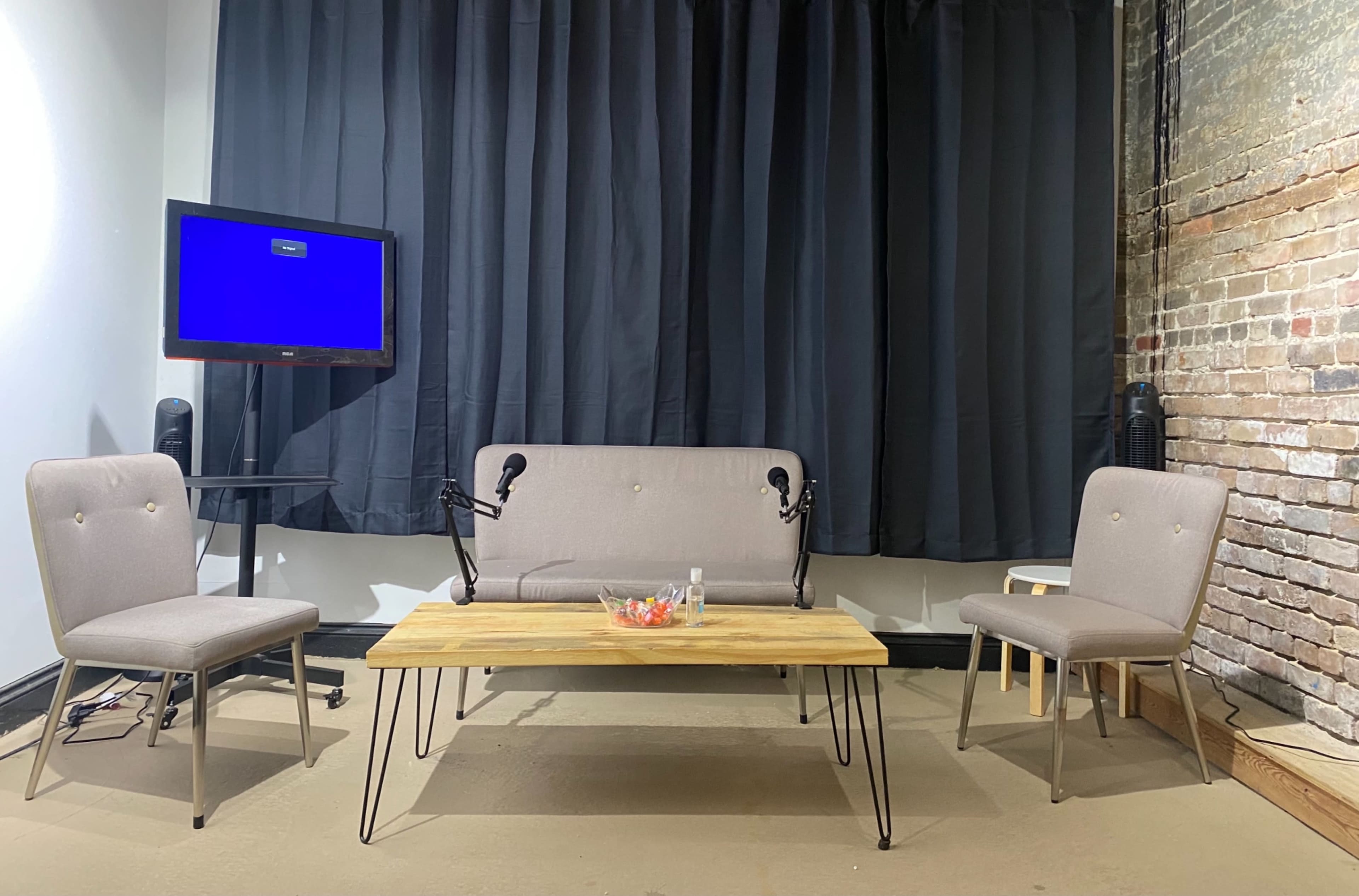 A small interview setup features a wooden table and two chairs, flanked by a couch, with a television displaying a blue screen in a brick-walled studio.