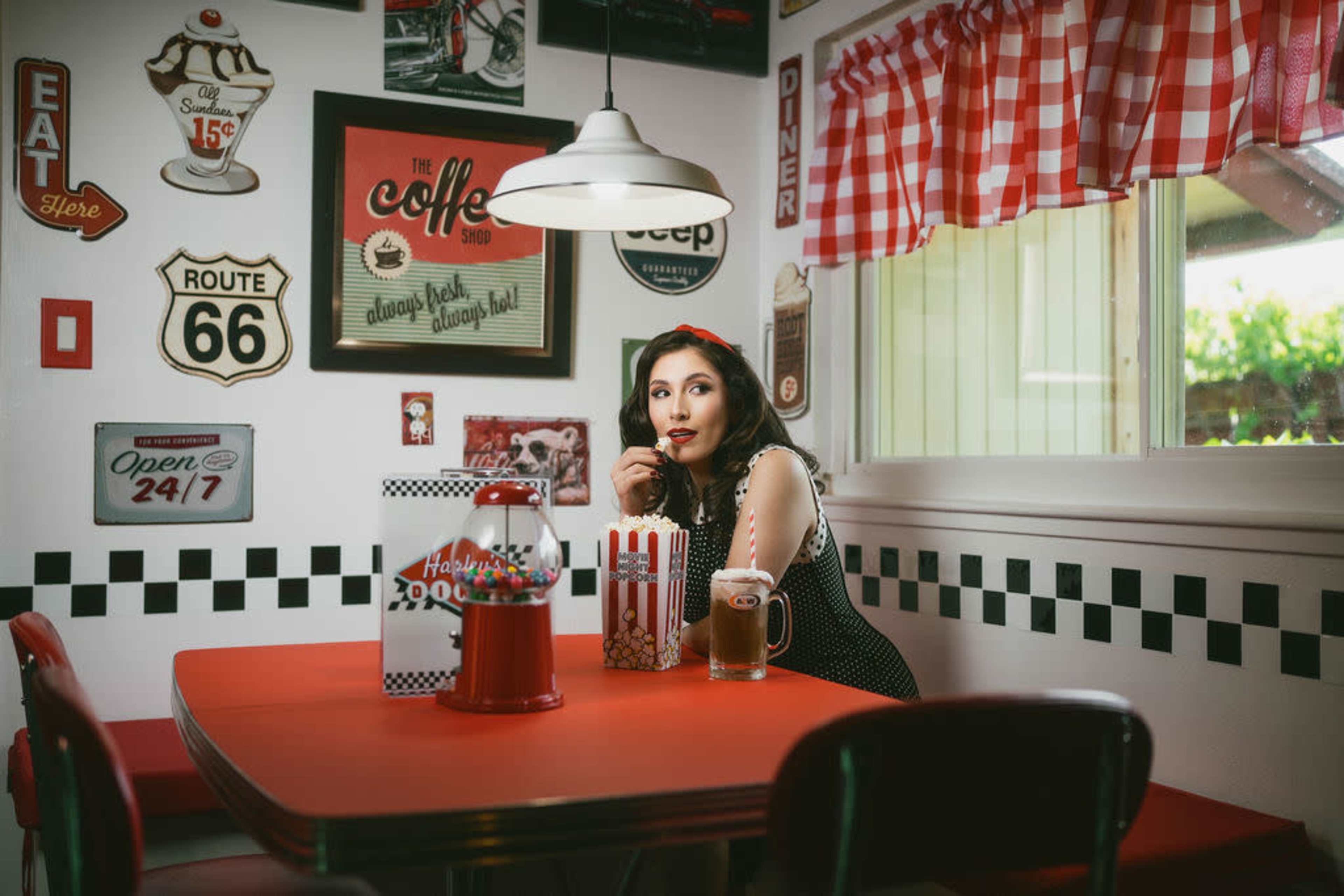 A woman sits at a red table in a retro diner, surrounded by vintage decor and a popcorn machine.