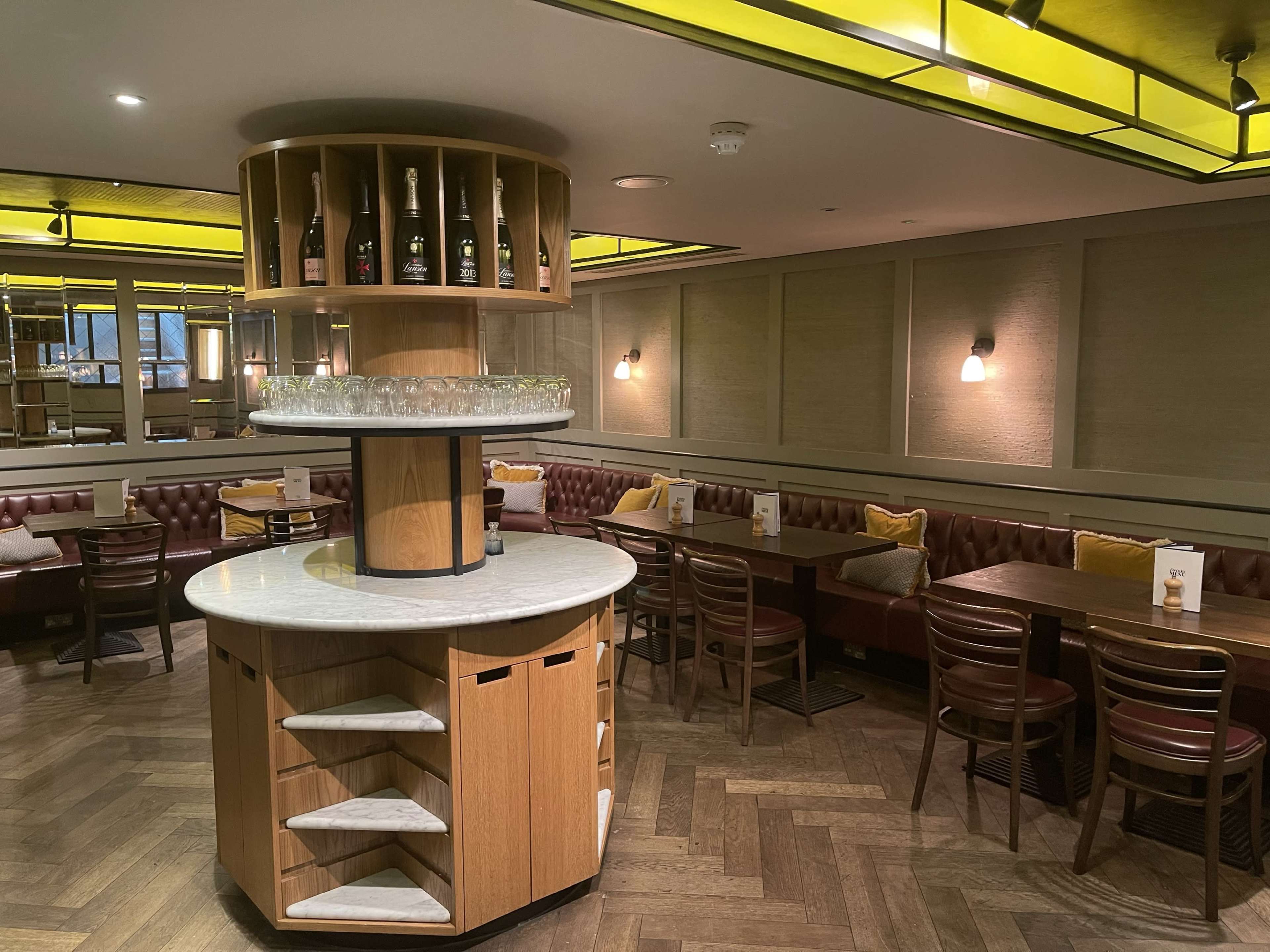 The image shows a modern restaurant interior featuring a central wooden display stand with shelves and bottles, surrounded by cozy seating areas with leather banquettes and wooden tables.