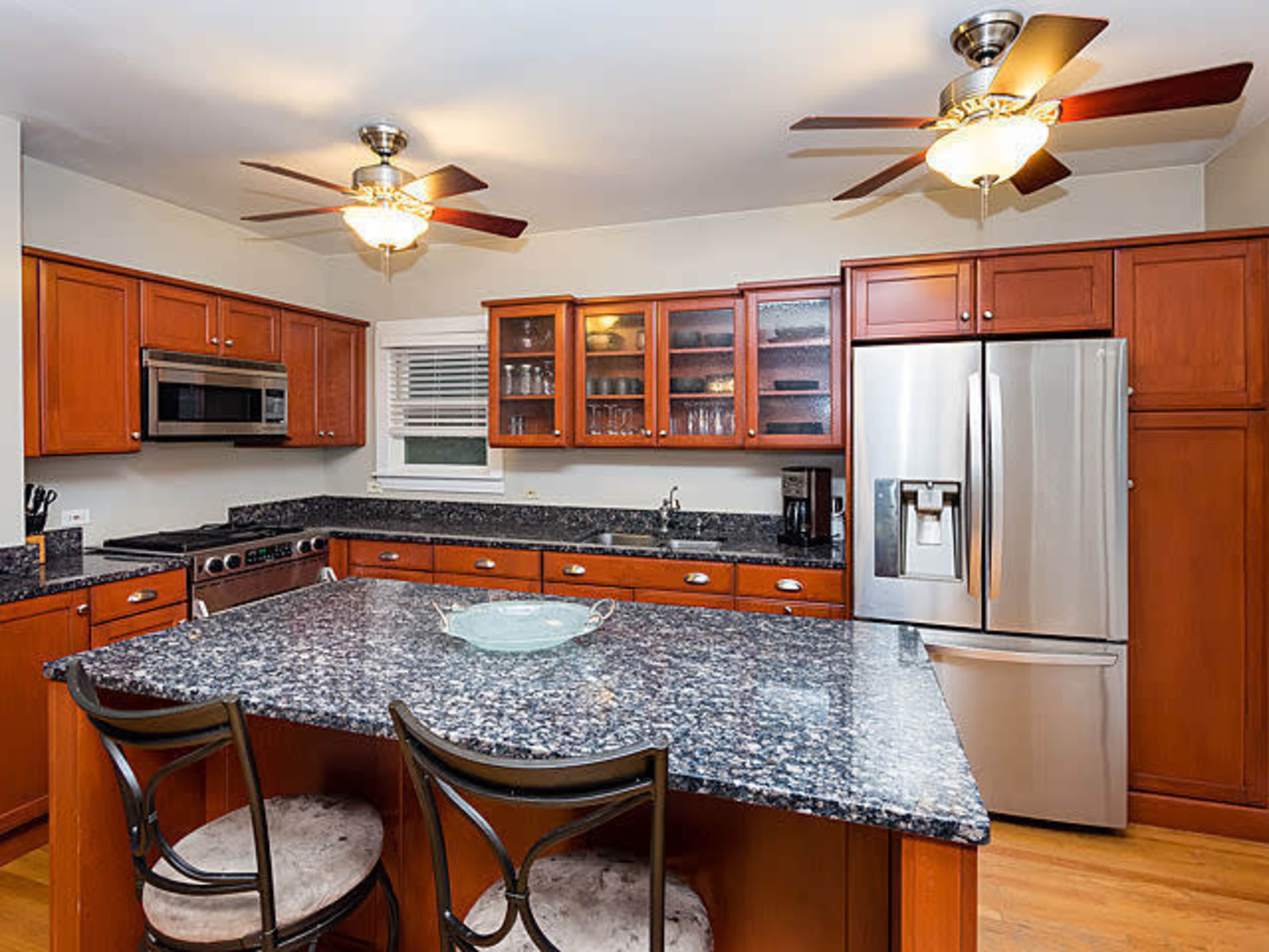 The image shows a kitchen with wooden cabinets, a granite countertop, and stainless steel appliances, featuring two ceiling fans and a large island with seating.