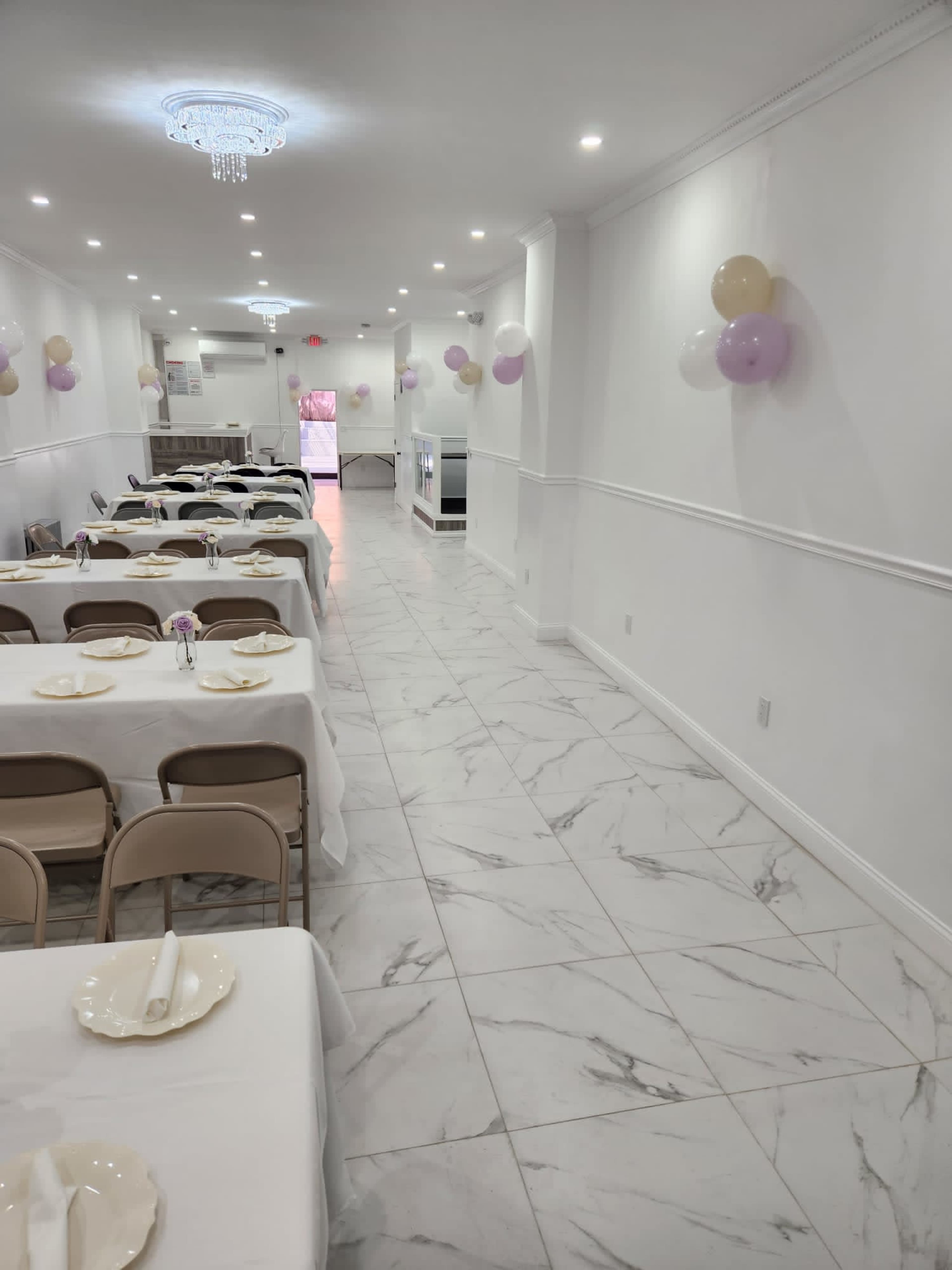 A long, narrow dining area with white tables covered in tablecloths, set with plates, and adorned with balloons along the walls.