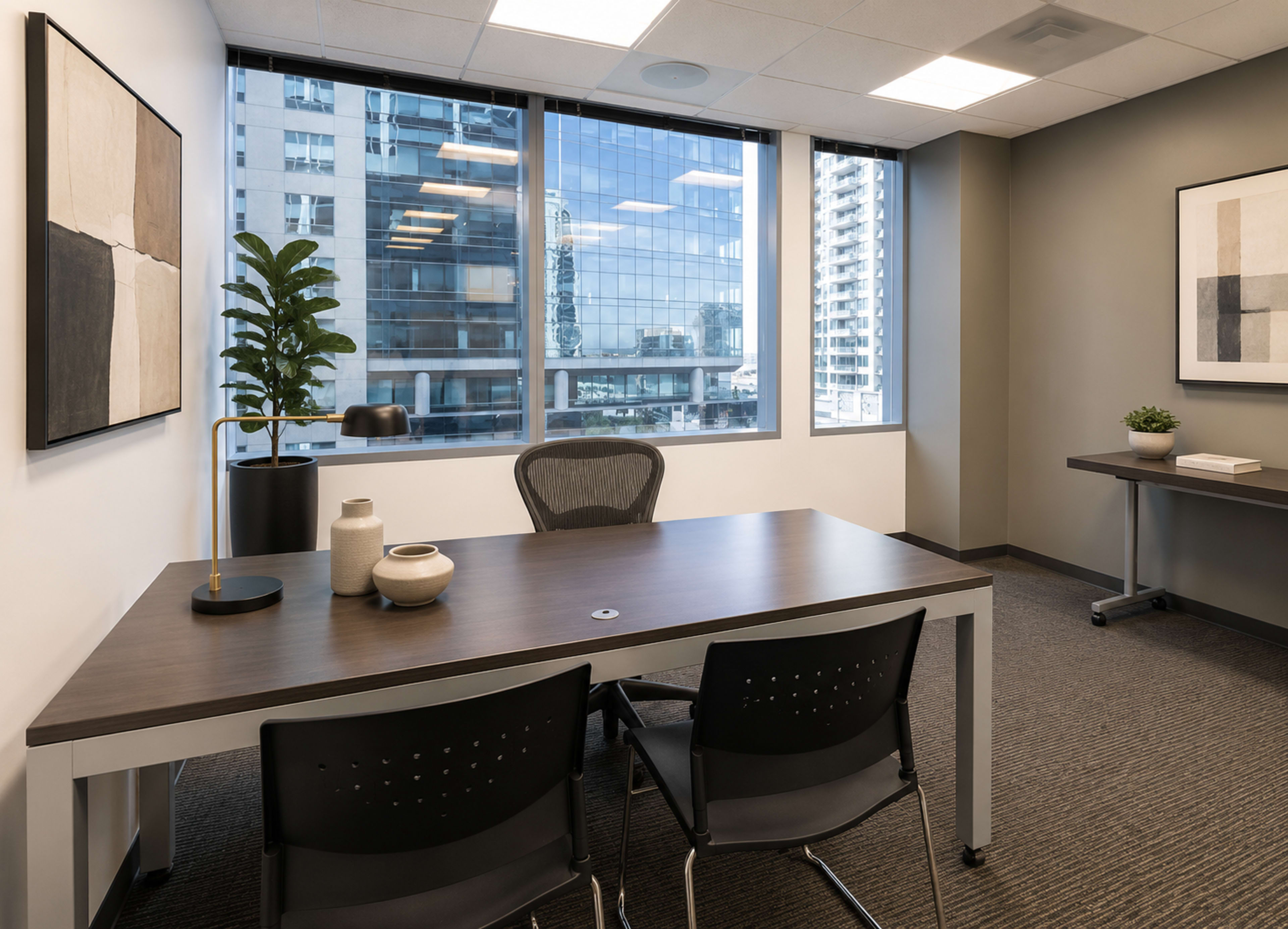 The image shows a modern office space featuring a wooden desk with two black chairs, large windows overlooking city buildings, and minimalistic decor on the walls.
