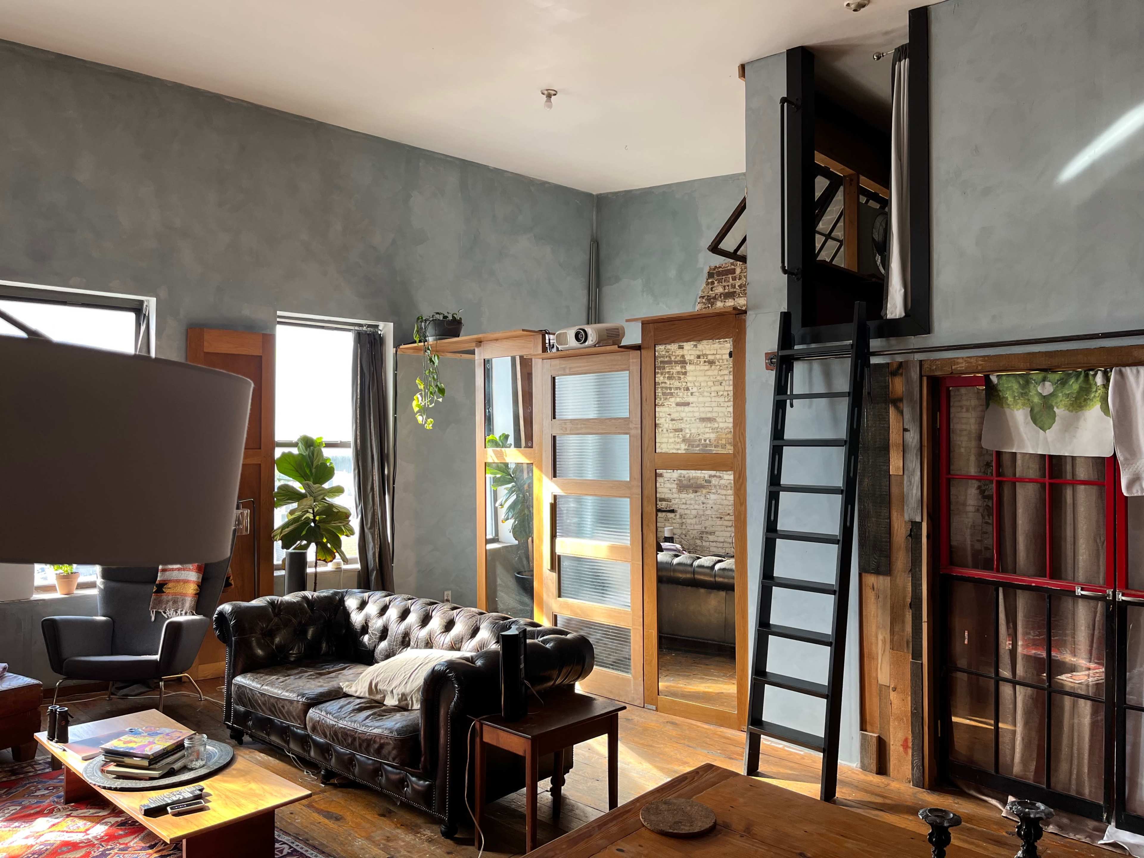 A modern, industrial-style living space featuring a leather sofa, a ladder leading to an upper level, and large windows that allow natural light to illuminate the room.