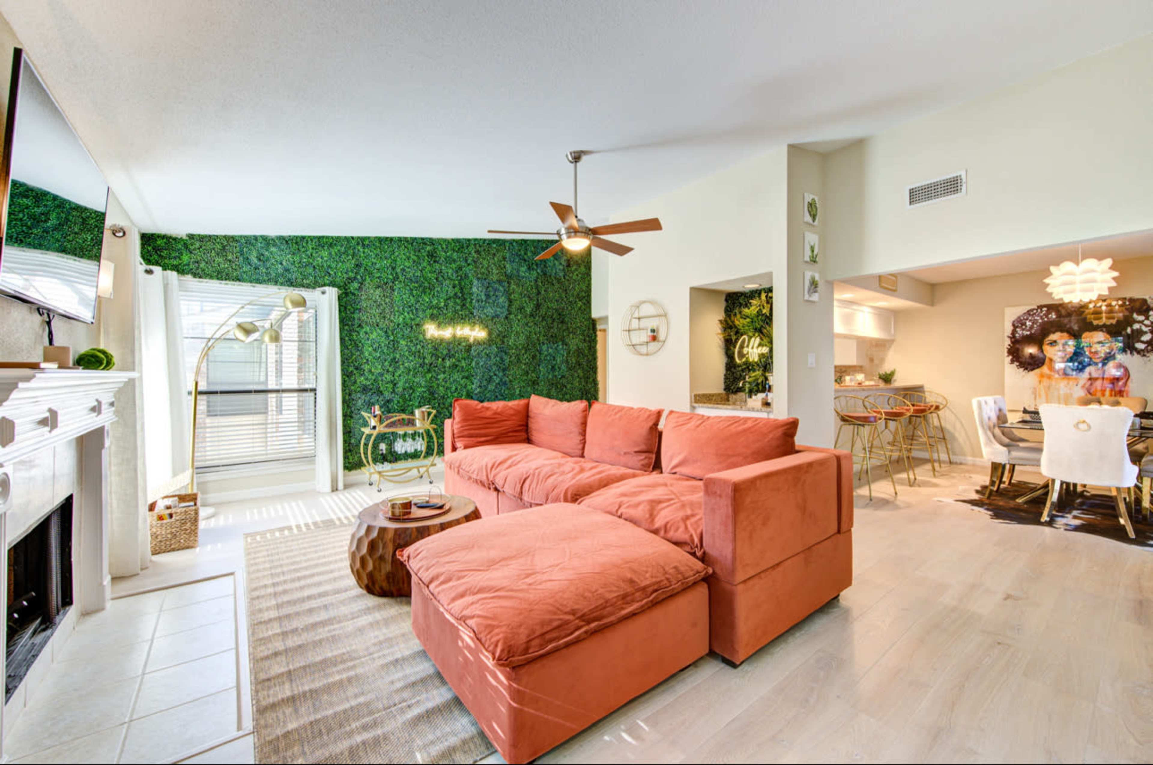 The image shows a modern living space featuring a large, orange sectional couch, a green accent wall, and a dining area with a round table and colorful artwork.