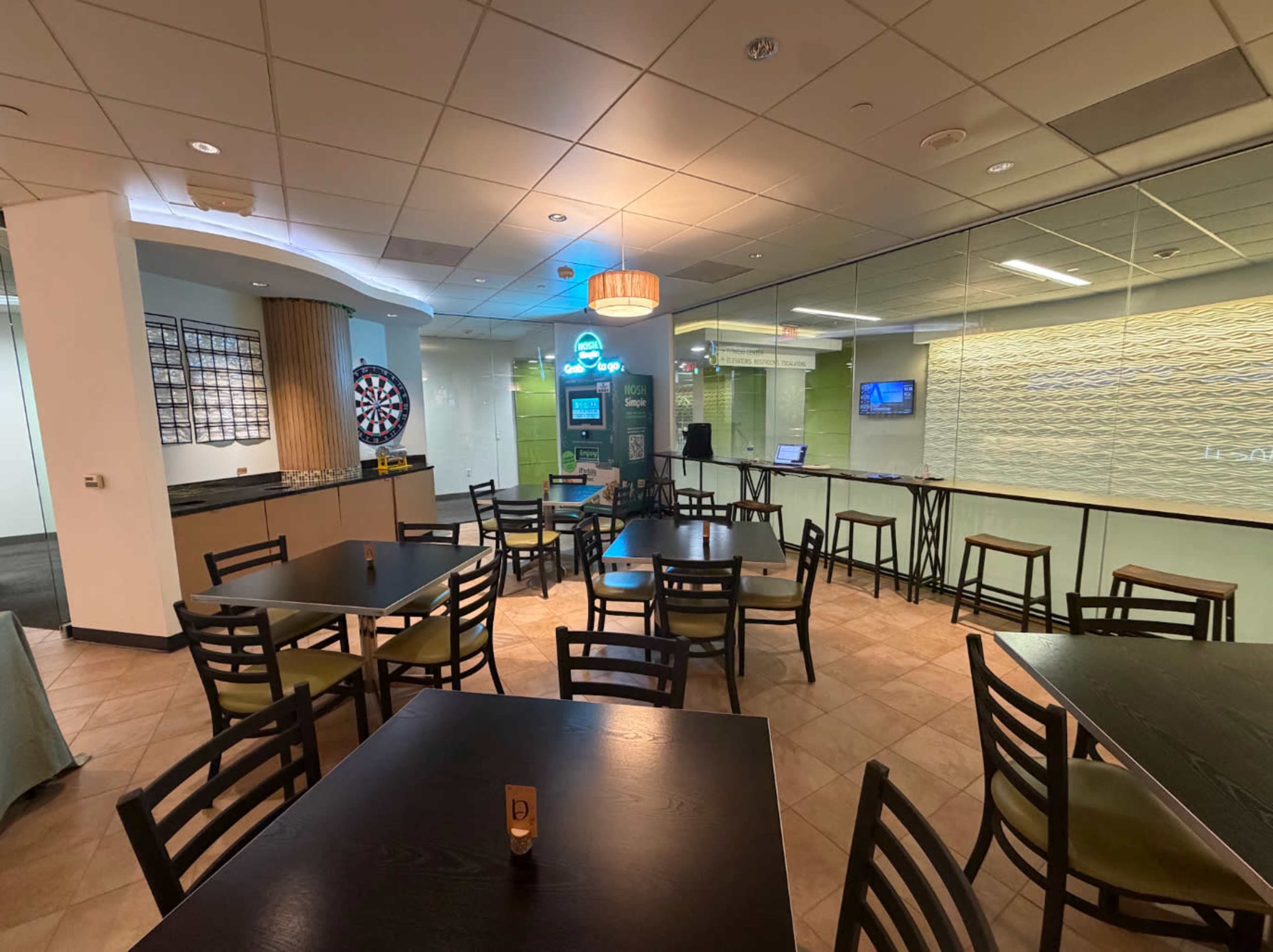 The image shows a modern dining area with tables and chairs, a dartboard, and a counter behind which there appears to be a service area or kitchen.