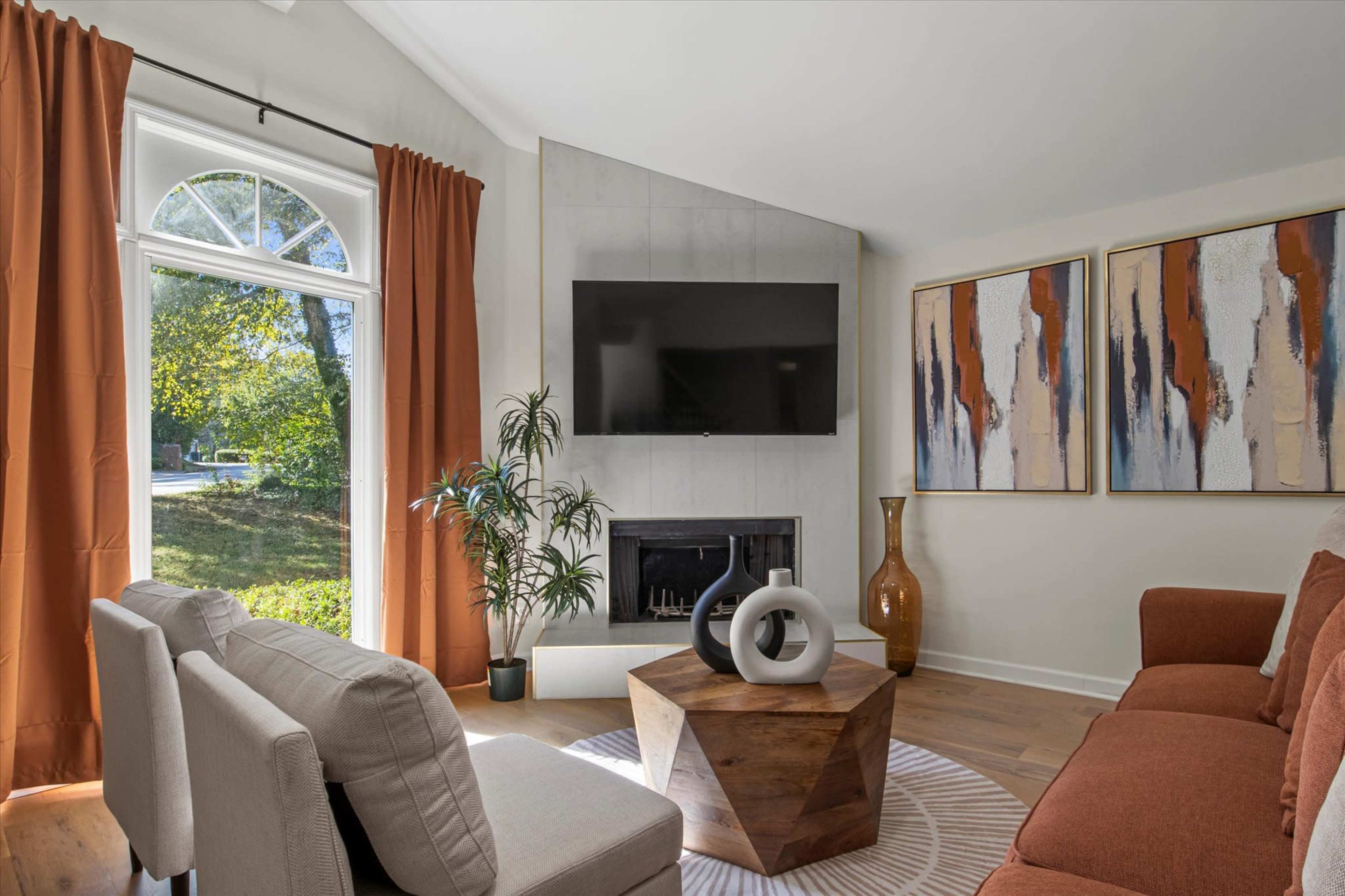 The living room features a large window with orange curtains, a wall-mounted television, a fireplace, and a coffee table surrounded by a sectional sofa and accent chairs.