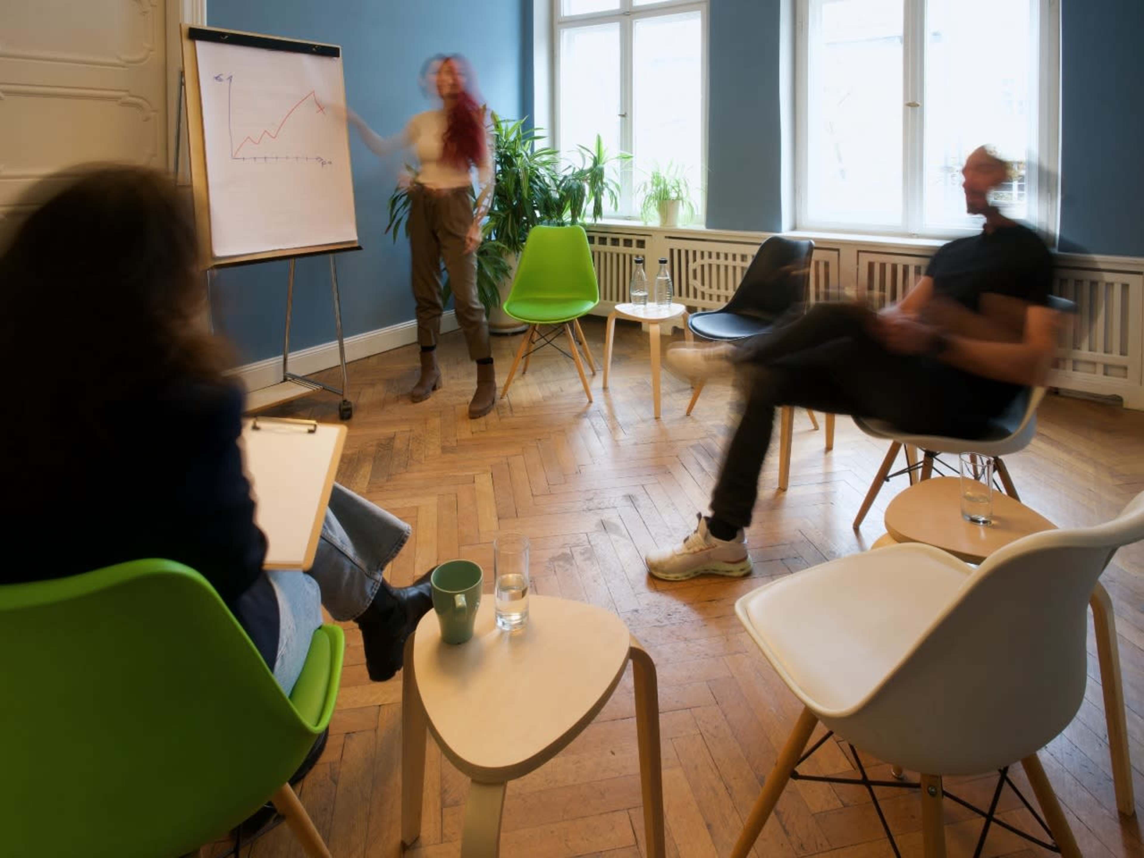 A woman with red hair stands in front of a flip chart displaying a line graph, while two people sit in chairs, one writing and the other leaning back.