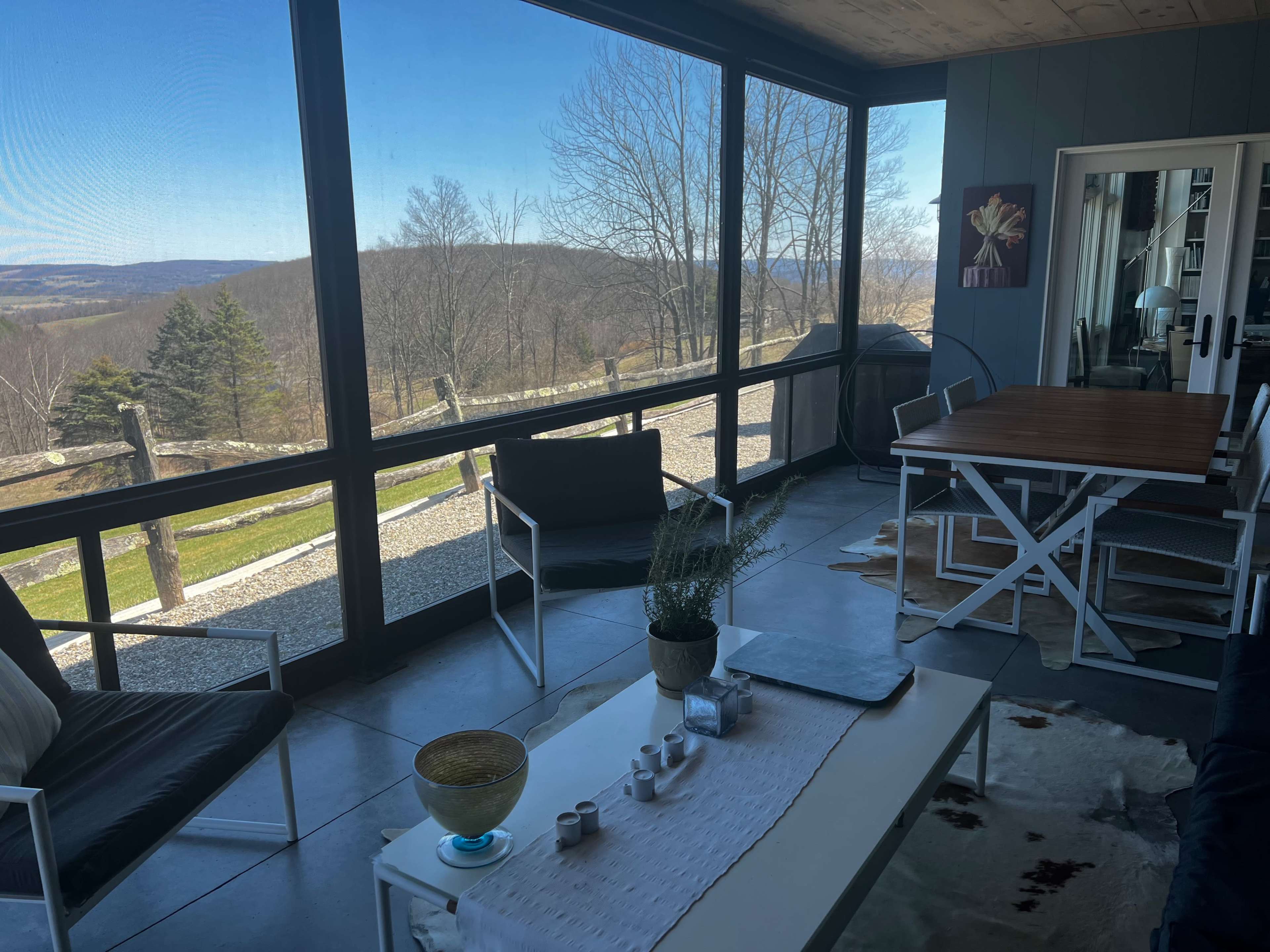 A sunlit screened porch features a dining table, seating area, and a view of rolling hills and trees through large windows.
