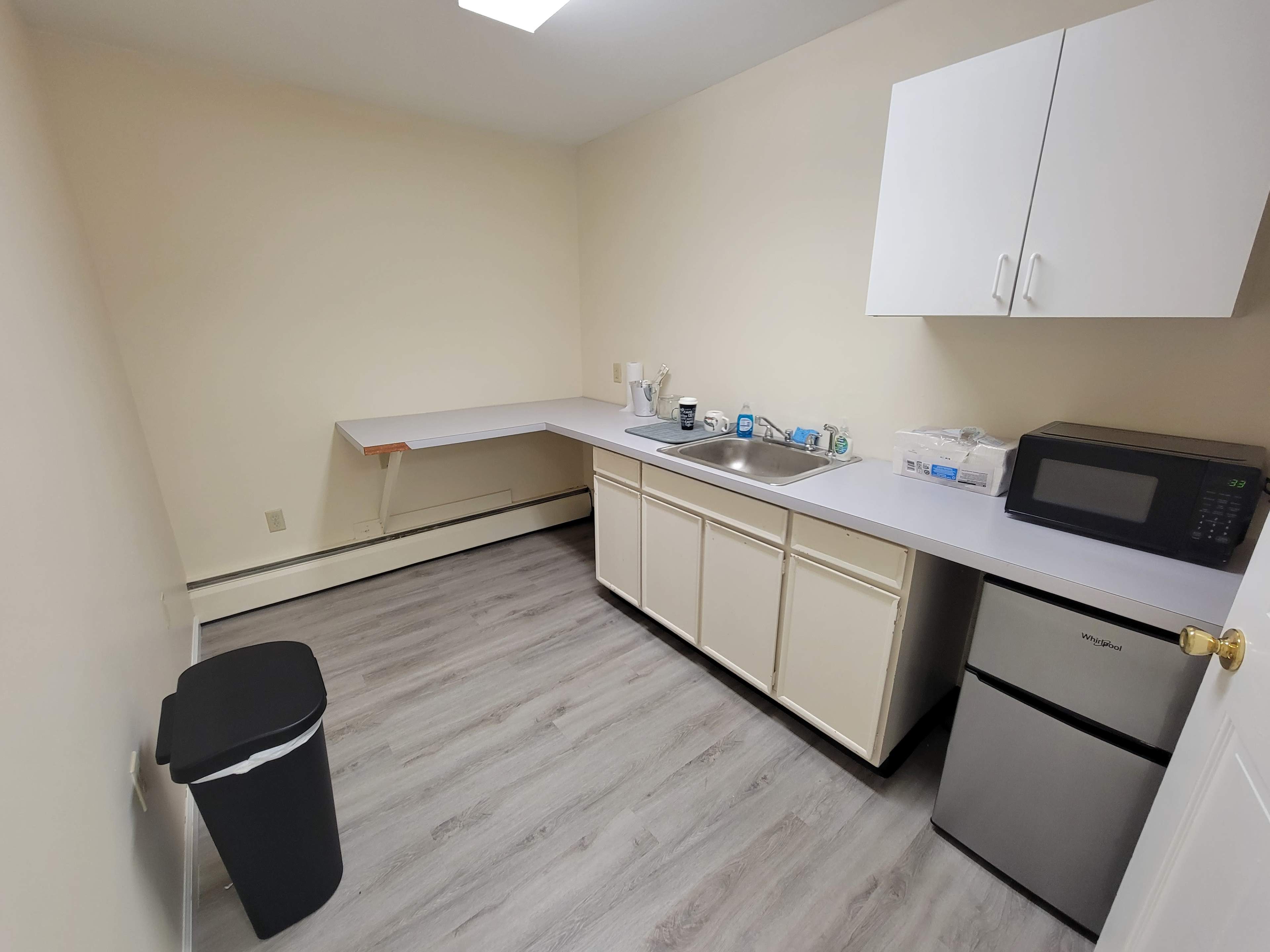 A small kitchen area with a sink, countertop, microwave, and a trash can, featuring white cabinets and light-colored flooring.