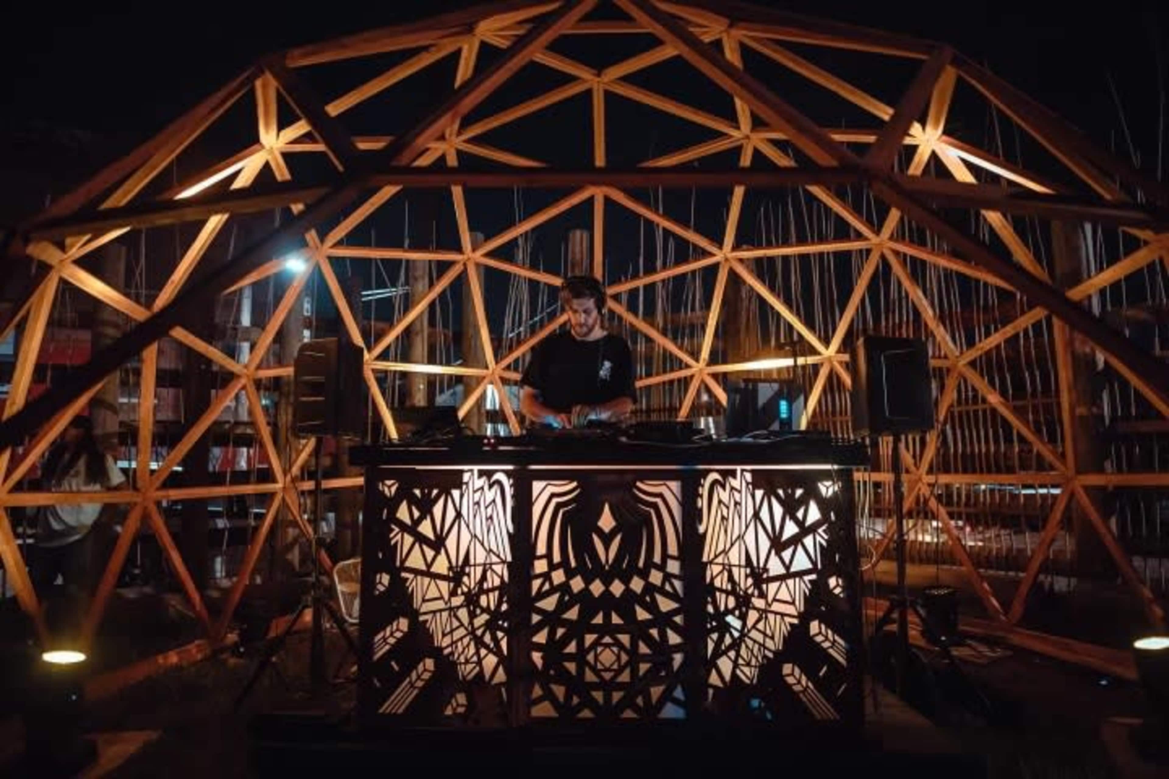A DJ performs inside a geometric wooden structure illuminated by stage lights.