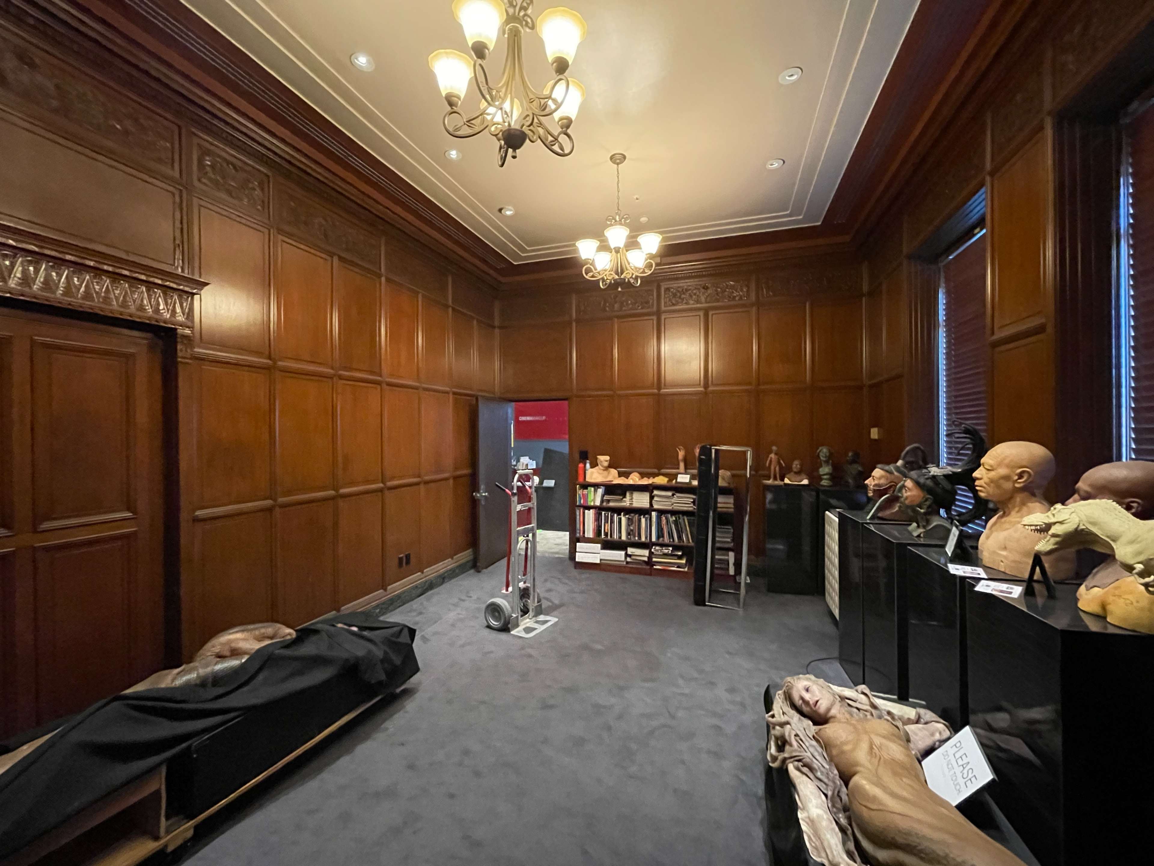 The image shows a dimly lit room with wooden paneling, featuring shelves of busts, a vacuum cleaner, and a partially visible display.