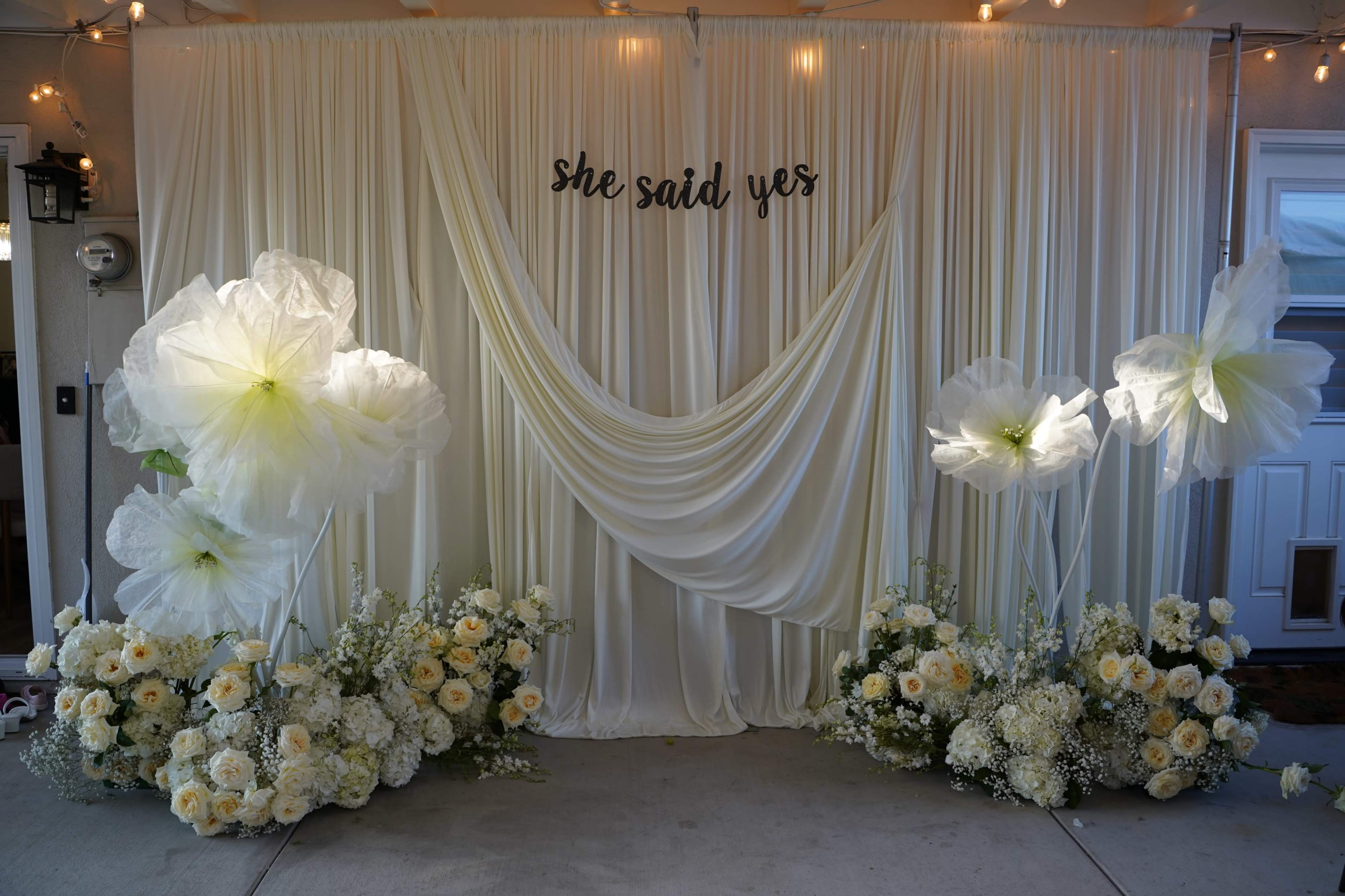A decorative backdrop featuring white fabric draping and floral arrangements of yellow and white flowers, with the phrase "she said yes" displayed prominently above.