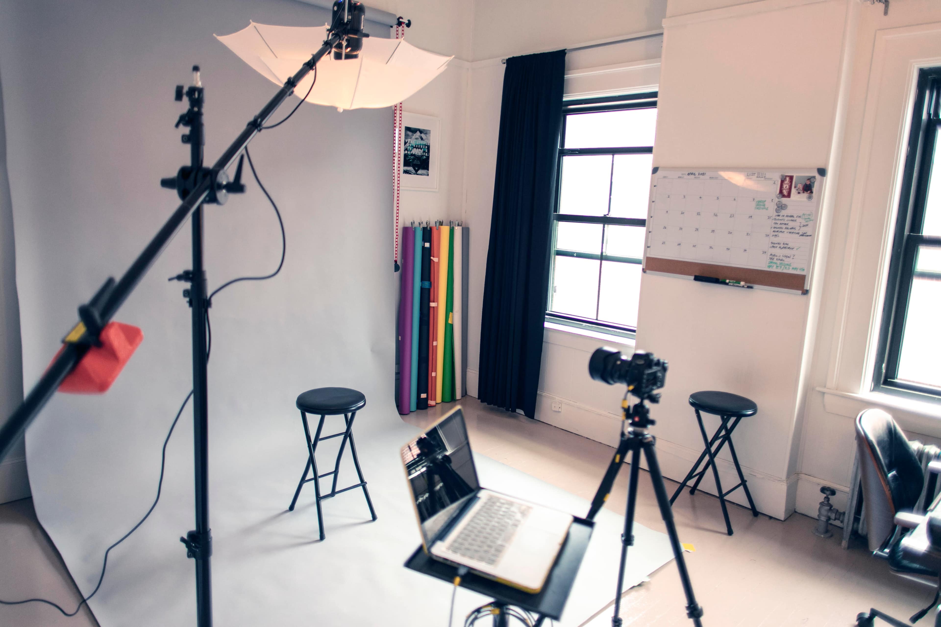 A photography studio setup includes a backdrop, lighting equipment, two stools, a camera on a tripod, and a laptop on a table.