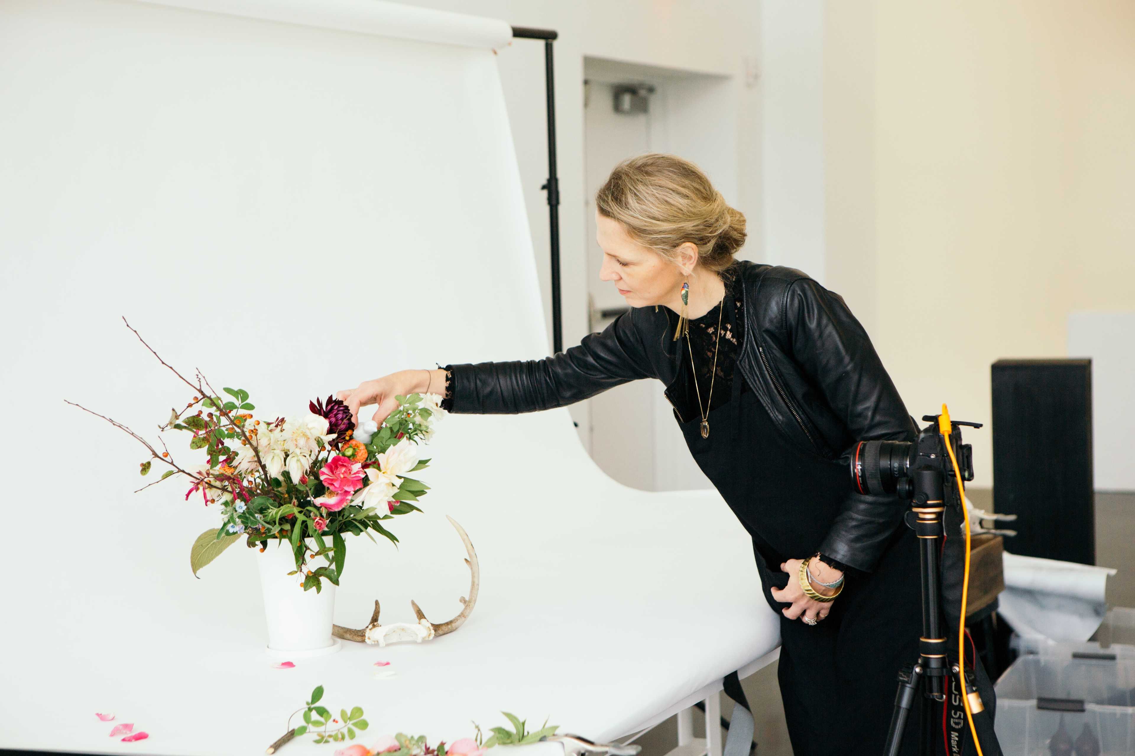 A woman arranges a flower bouquet next to an antler on a white backdrop in a studio setting.
