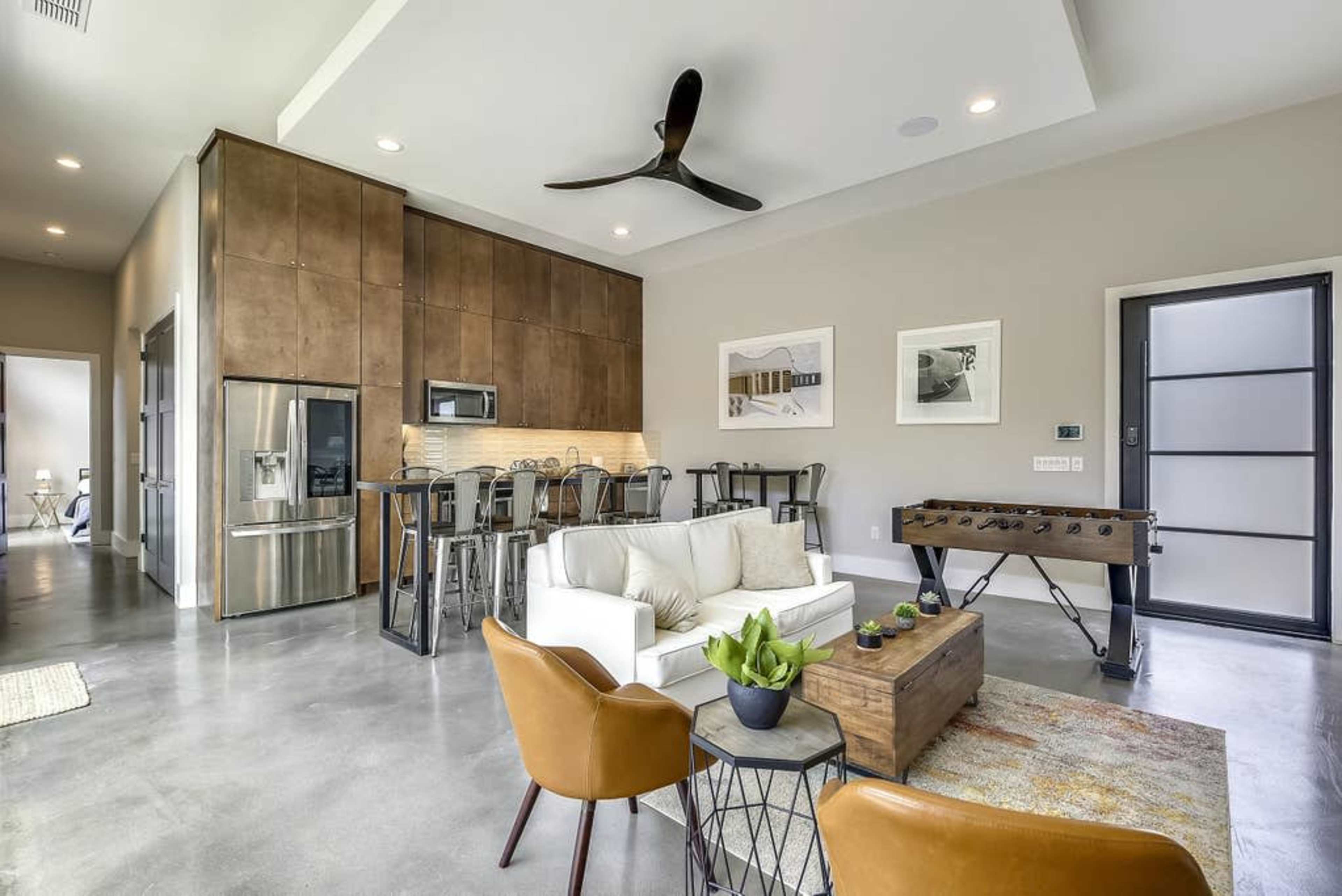 A modern living space with a light-colored sofa, a wooden coffee table, and a foosball table, adjacent to a kitchen with bar seating and stainless steel appliances.