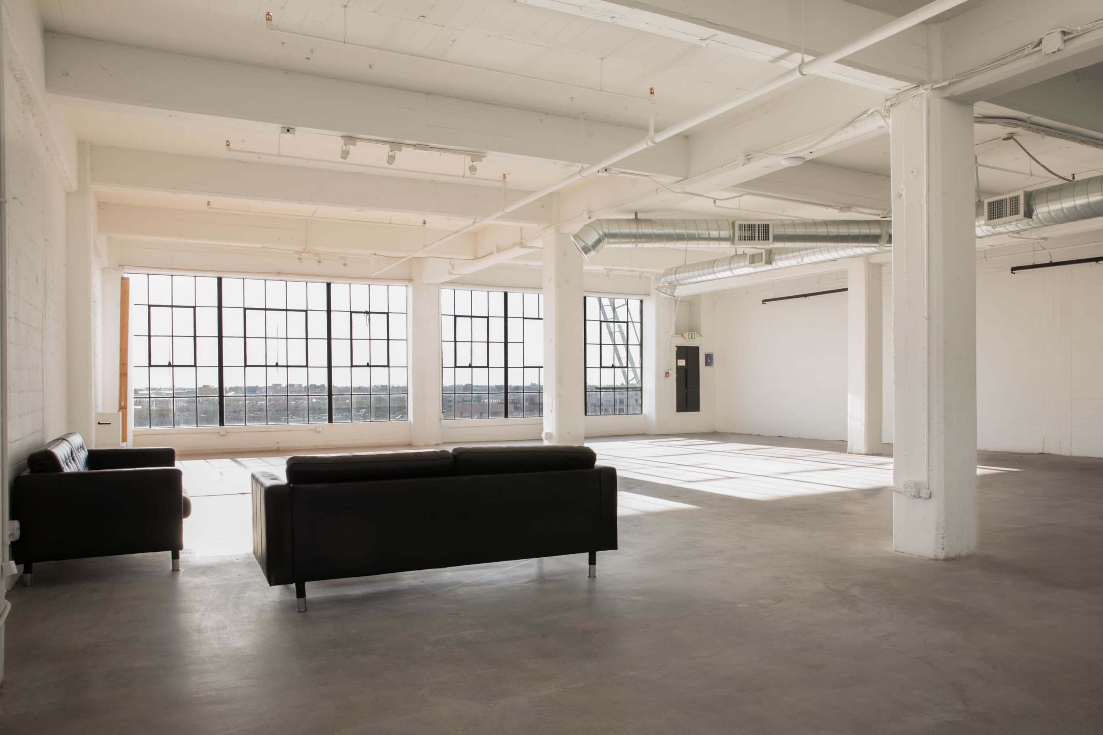 A spacious, bright industrial-style room features large windows, high ceilings, and a black couch in the foreground.