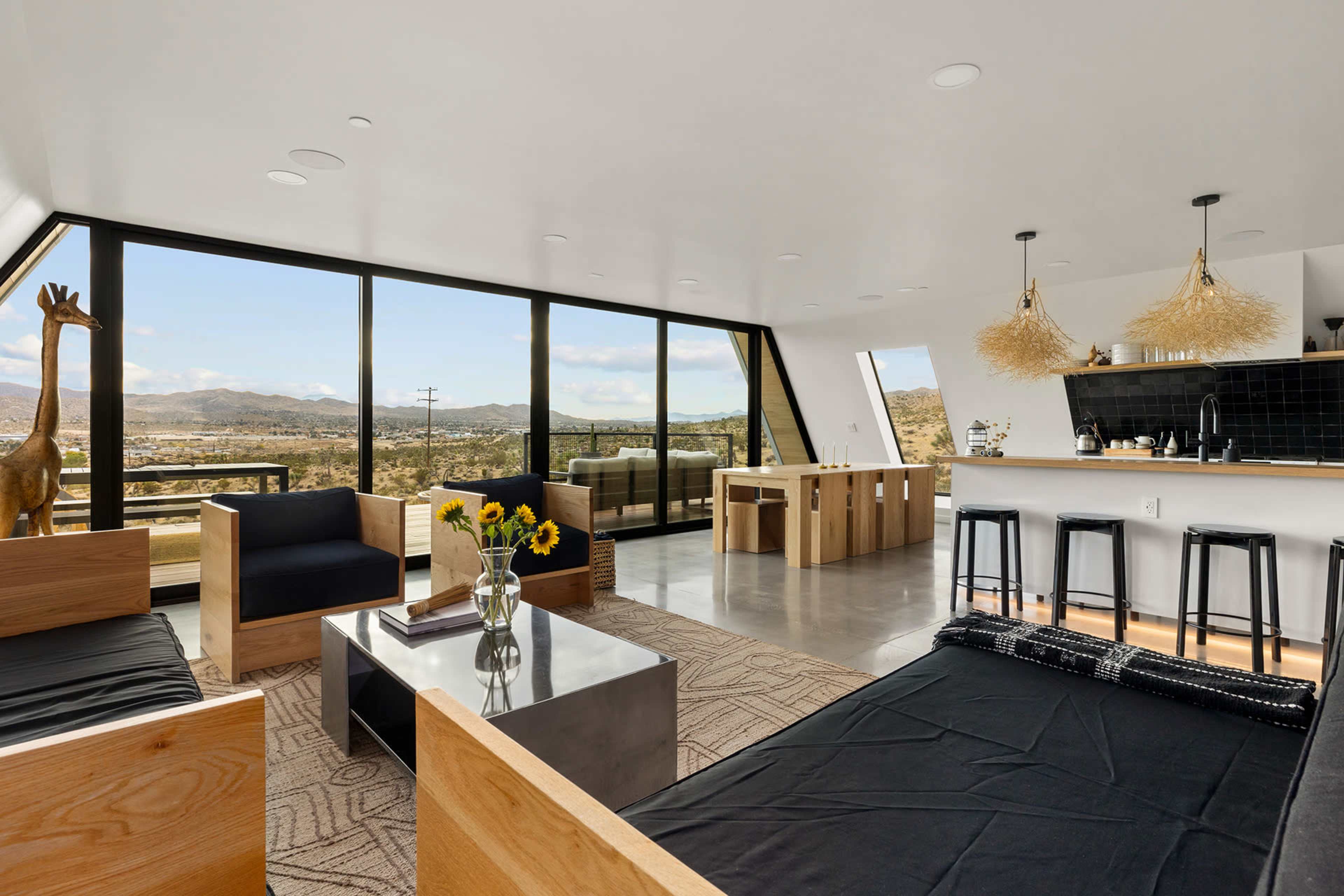 A modern living space features large windows offering a view of the landscape, with wooden furniture, a black and white decor, and a kitchen bar with stools.