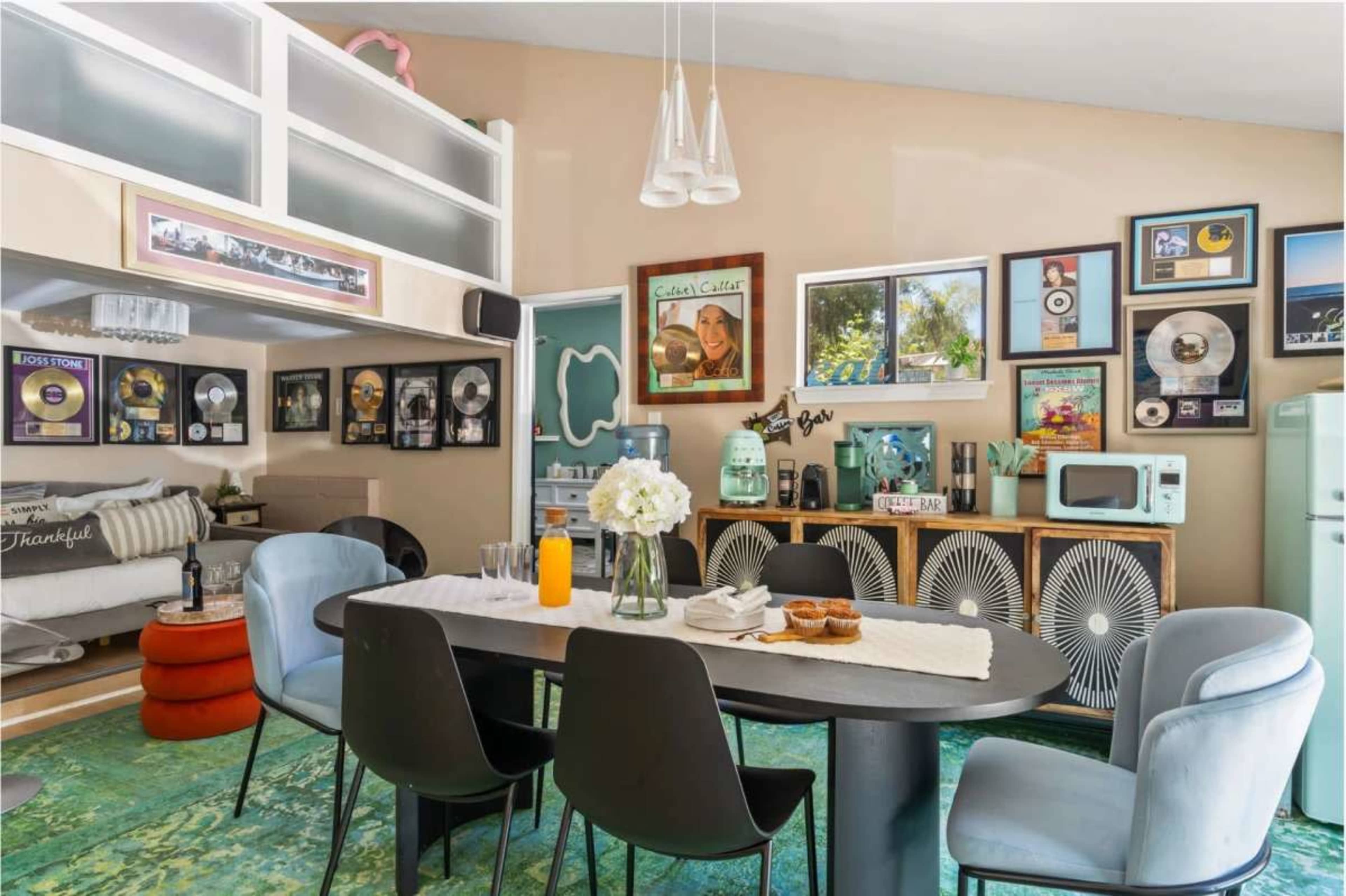 The image shows a modern dining area with a long table and black chairs, surrounded by framed music awards and decorative items on the walls.
