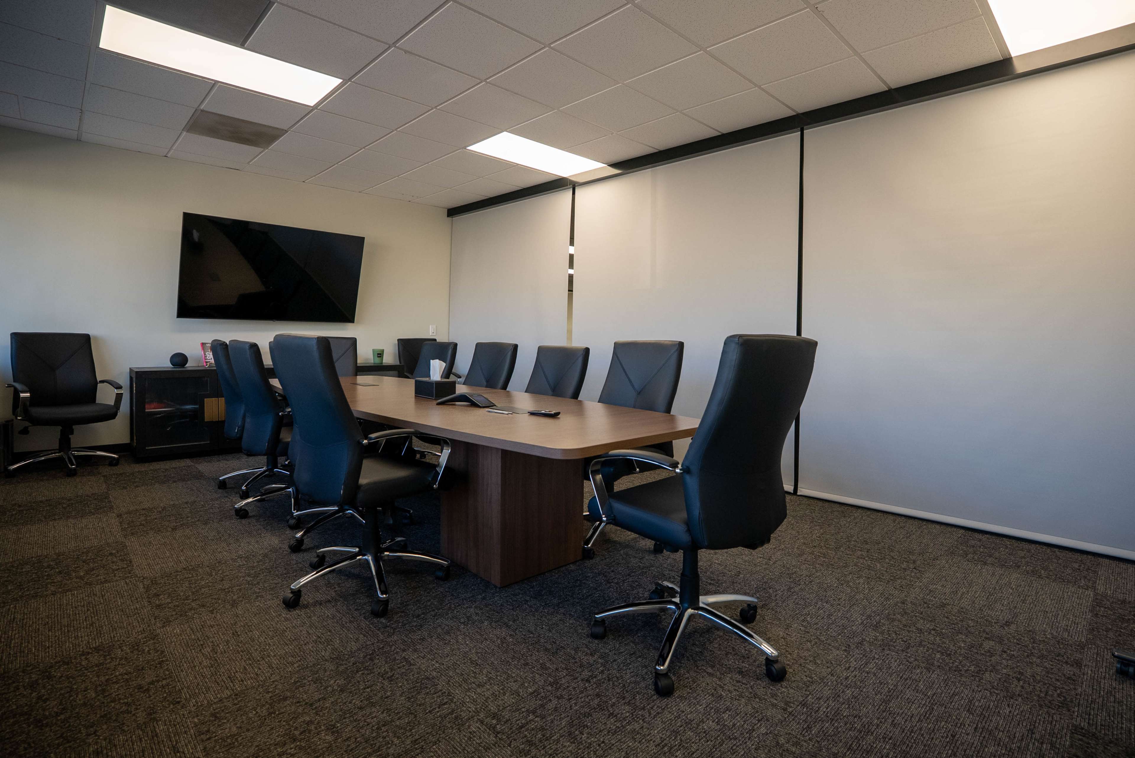 A spacious conference room features a long wooden table surrounded by black leather chairs, with a large television mounted on the wall.