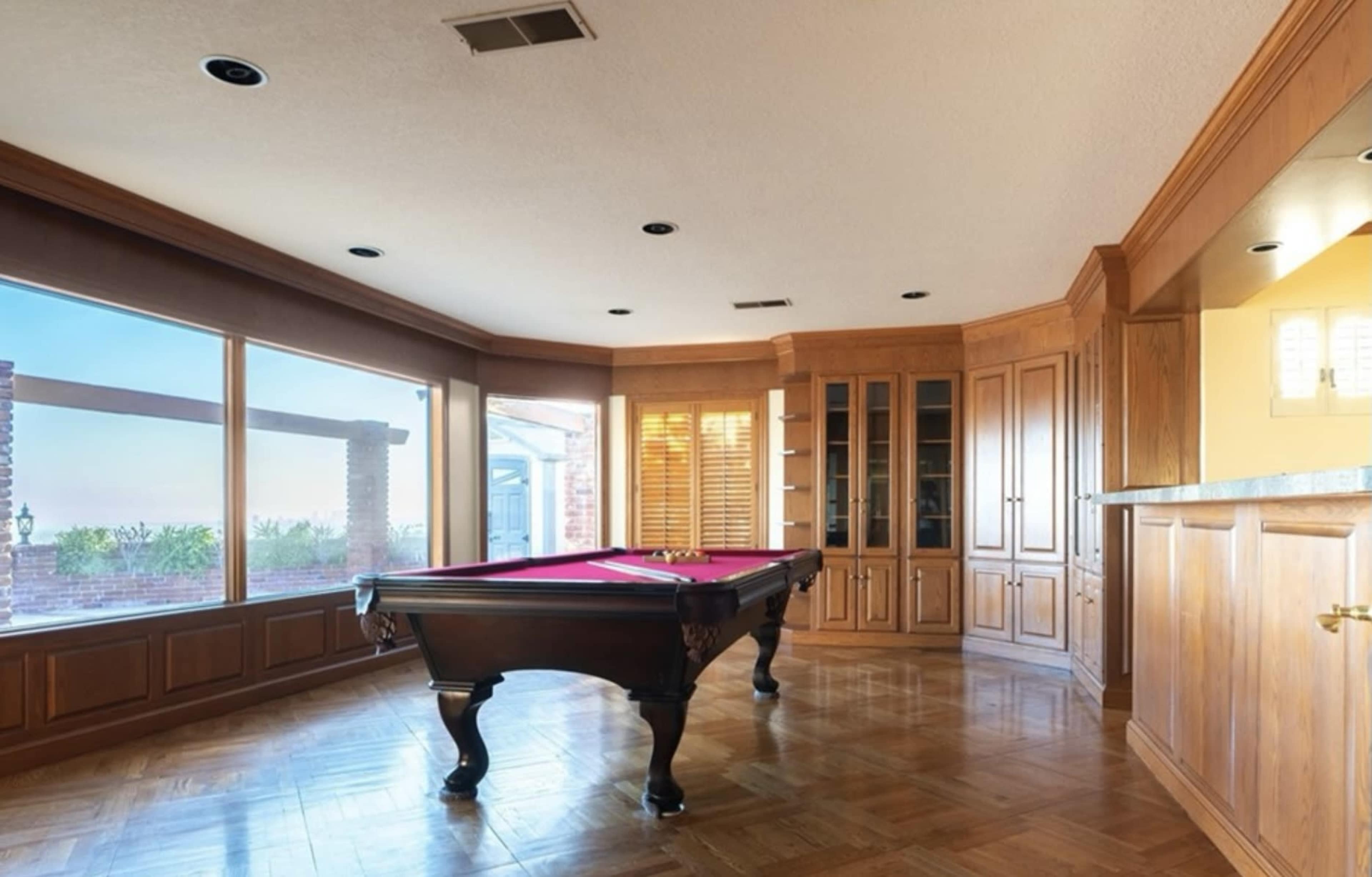 A wooden-paneled game room with a red felt pool table and large windows overlooking an outdoor view.