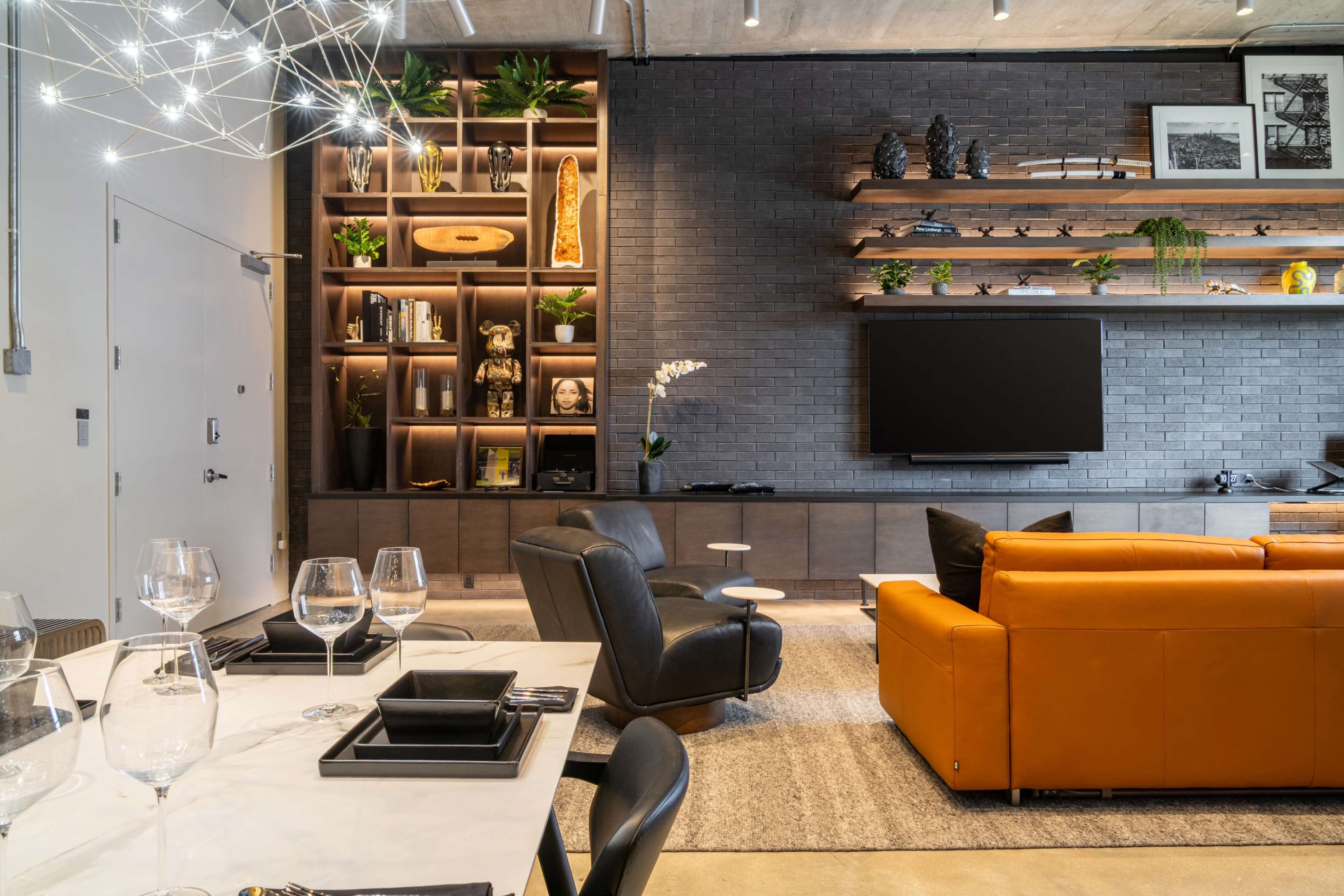 The image shows a modern living space with a sleek design, featuring a large orange sofa, a wall-mounted television, and a decorative shelf filled with plants and various objects.