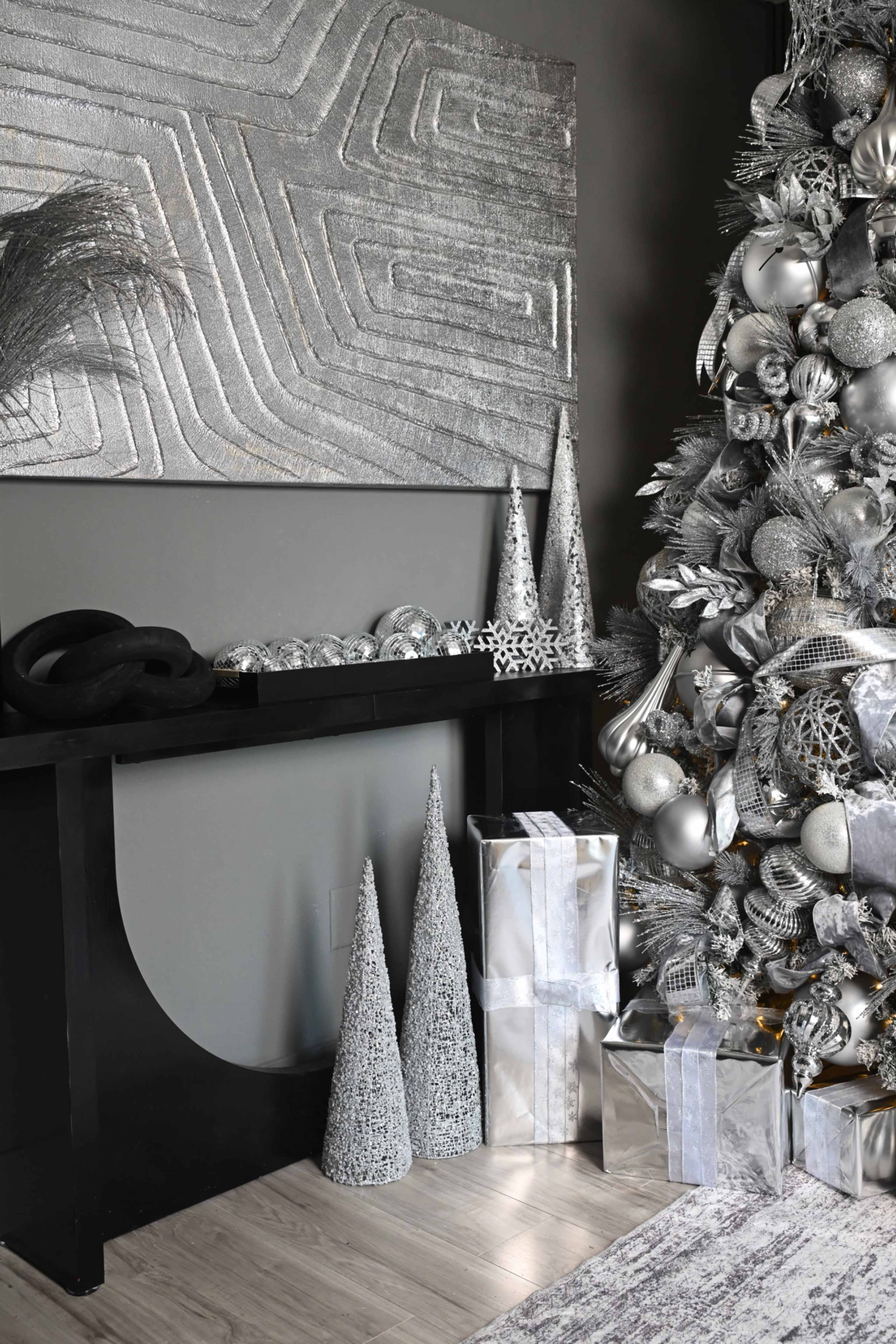 A decorated Christmas tree with silver ornaments stands next to a black console table adorned with metallic trees and wrapped gifts.
