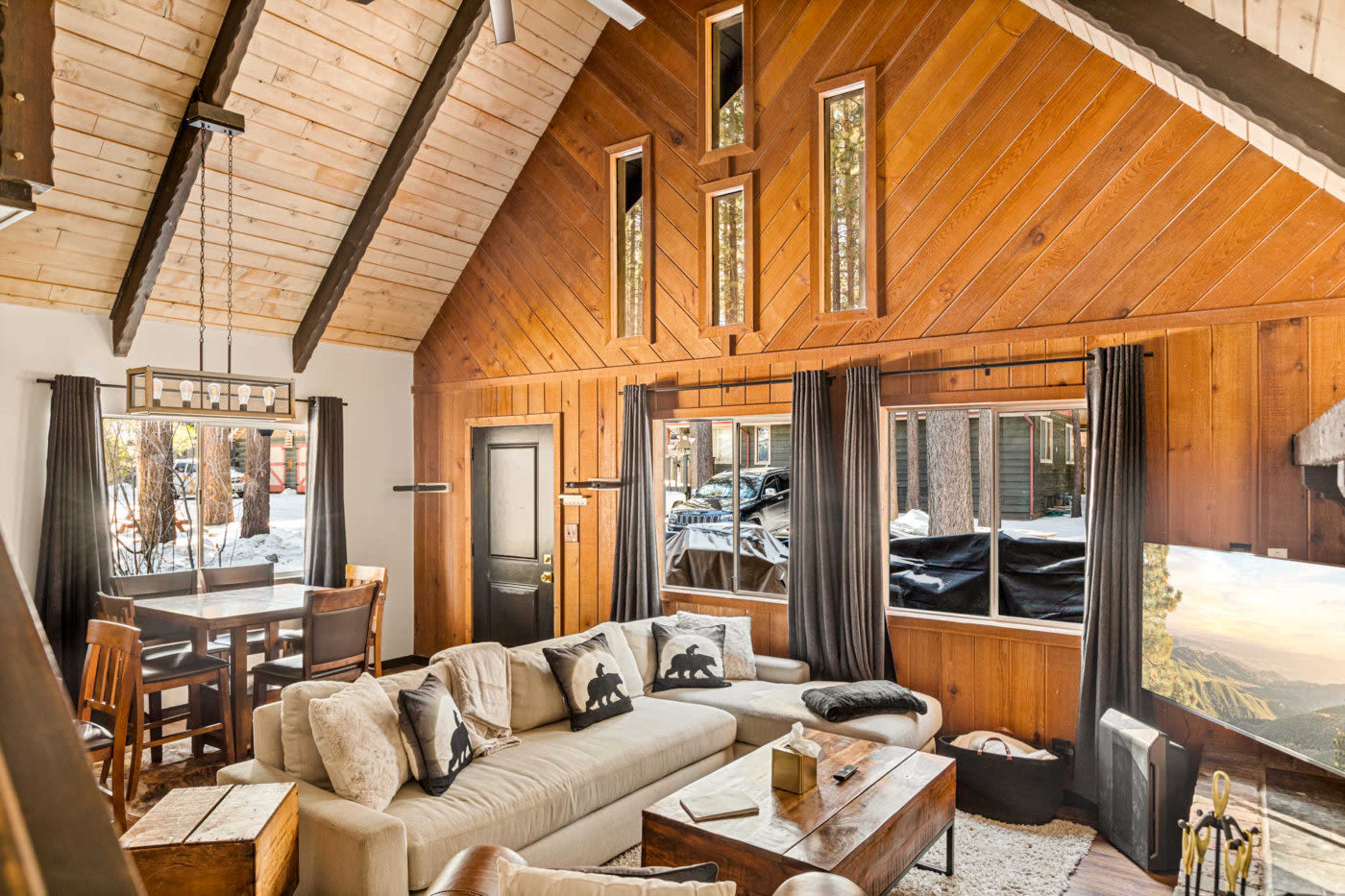 The living room features a large sectional sofa, wood paneling, high ceilings, and tall windows that provide a view of the snowy outdoors.