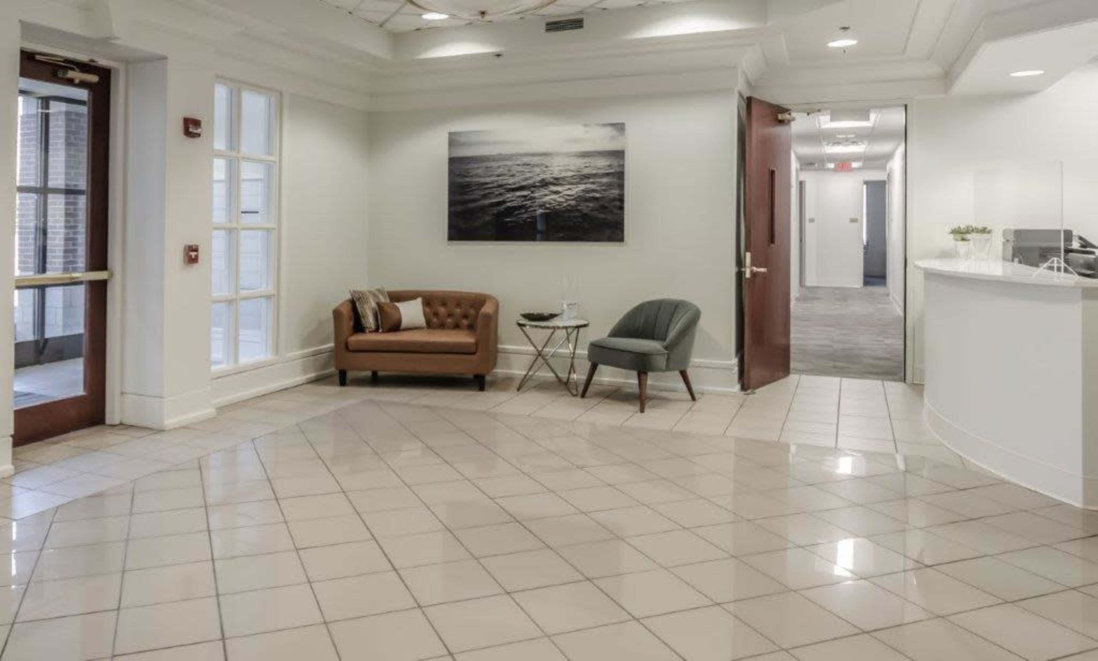A modern reception area featuring a brown leather couch, a green chair, and a white reception desk with gray flooring and a large photograph of the ocean on the wall.