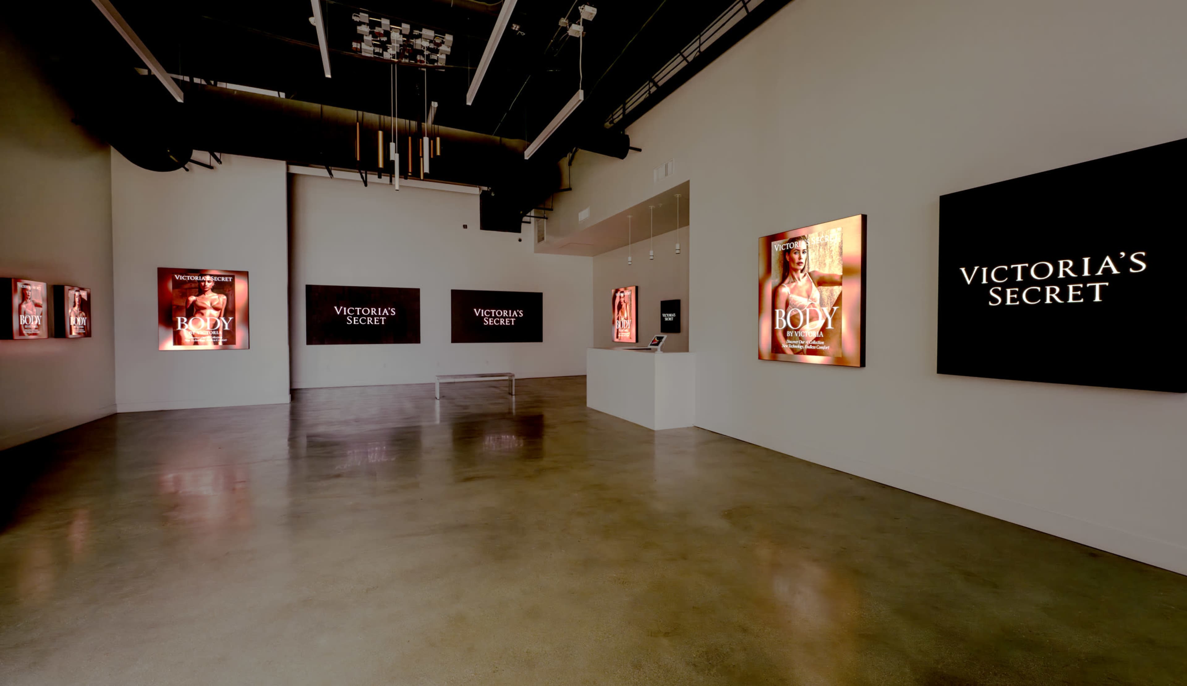 The image shows an exhibition space featuring multiple illuminated displays of Victoria's Secret advertisements on white walls and a central display unit.