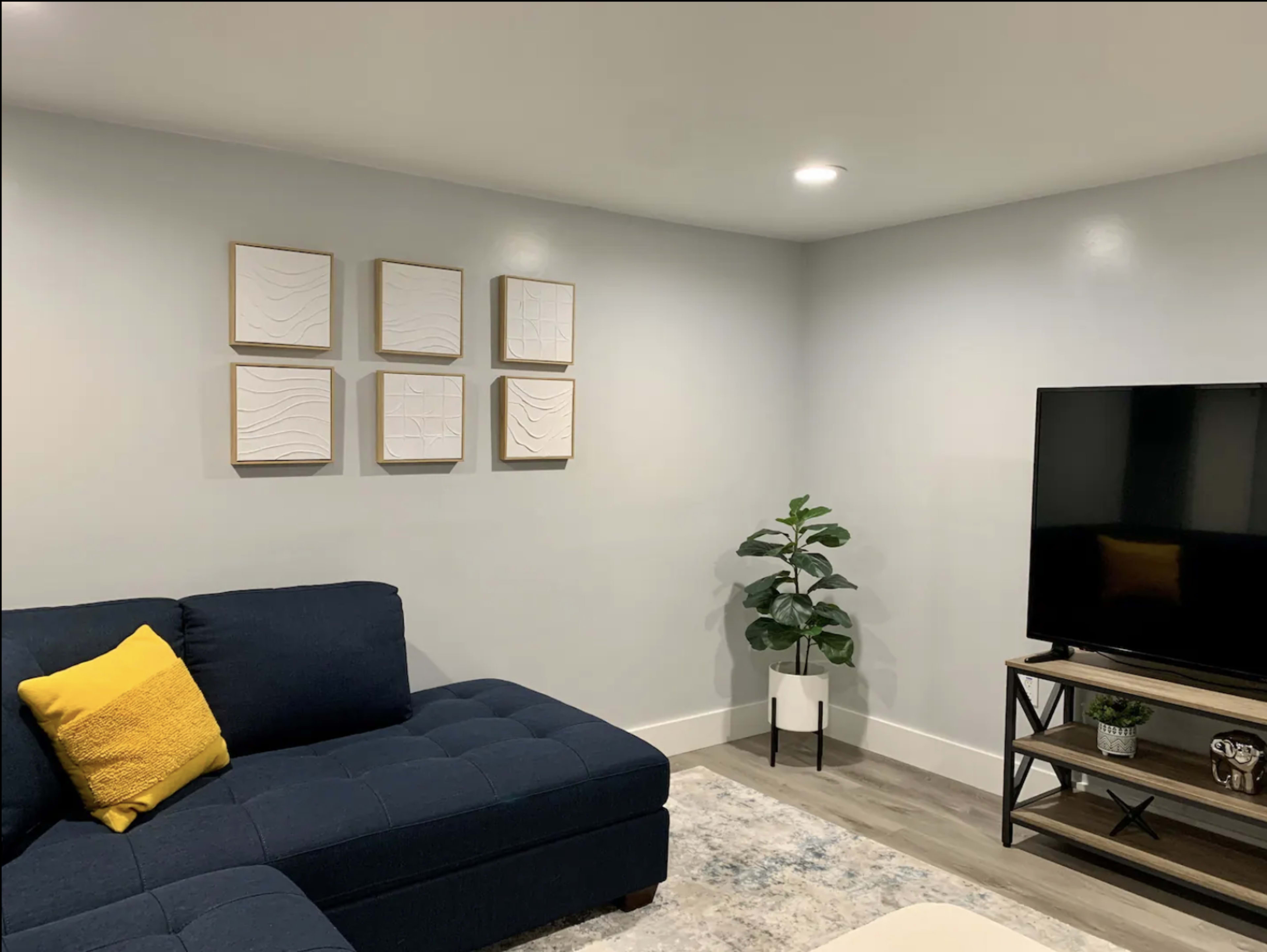 A modern living room with a blue sectional sofa, framed abstract wall art, a television on a stand, and a potted plant in the corner.