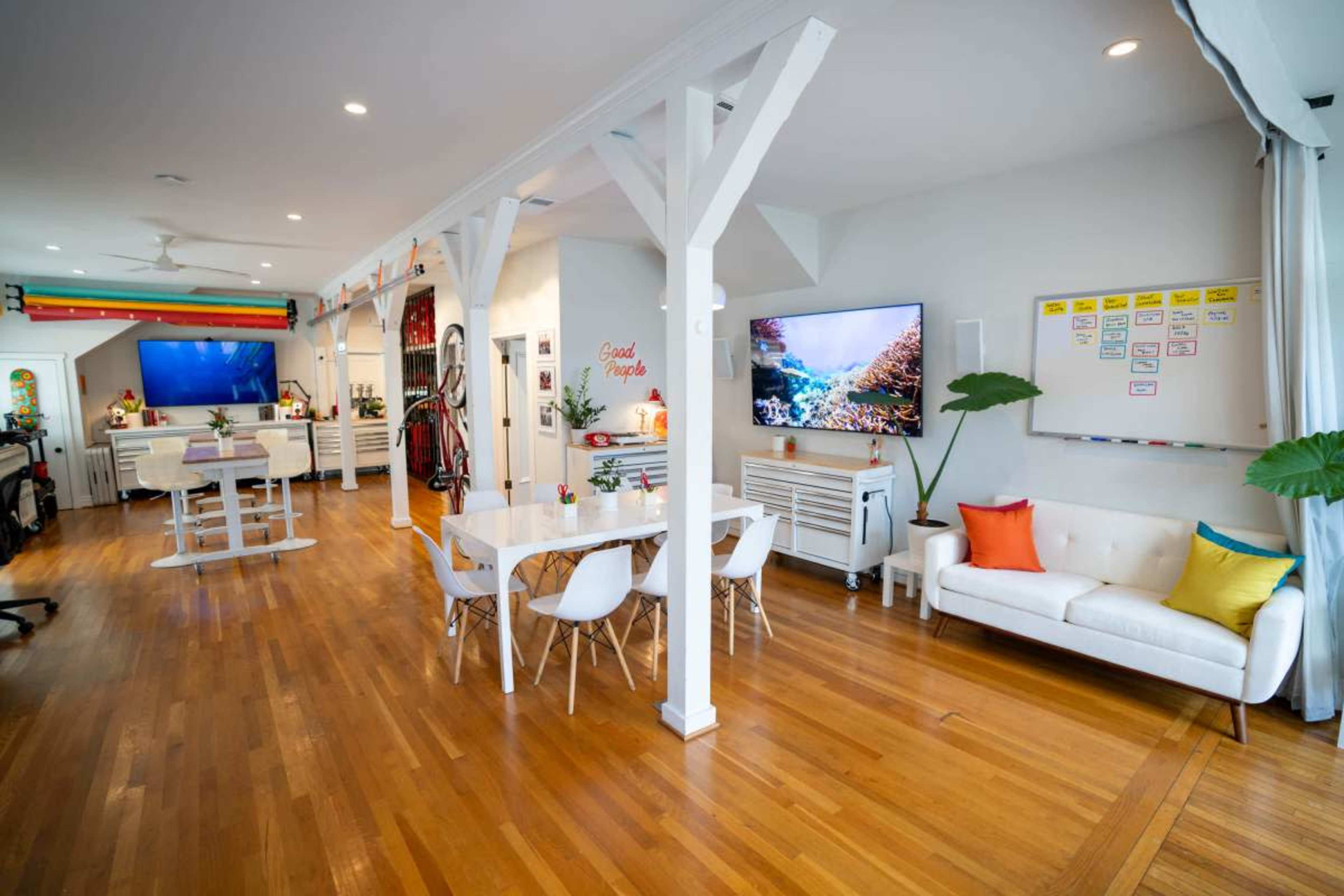 The image shows a modern, open-concept living space with a dining table, white furniture, and several televisions mounted on the walls.