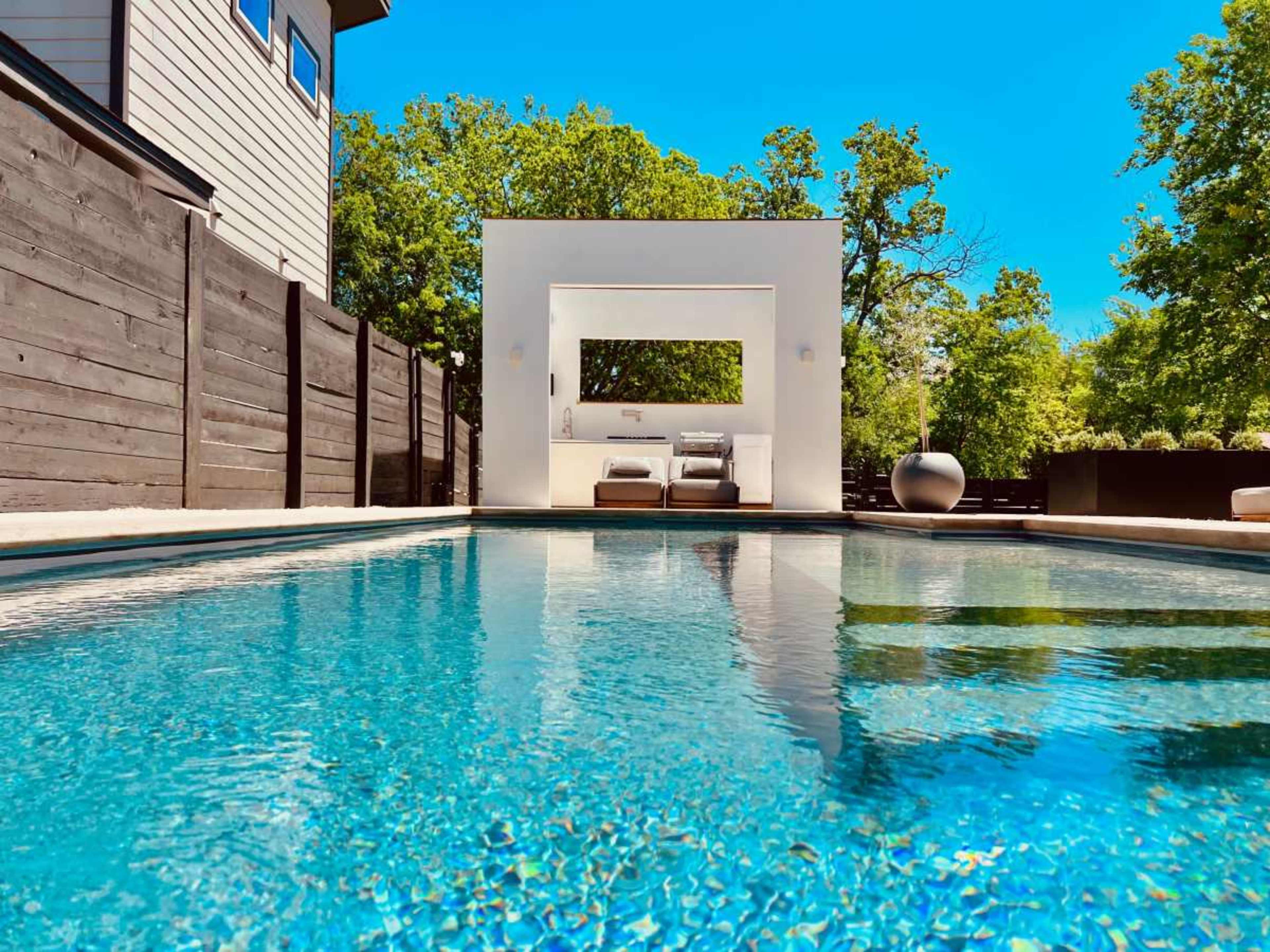 A modern outdoor pool reflects the clear blue sky, with a white seating area and lush greenery in the background.