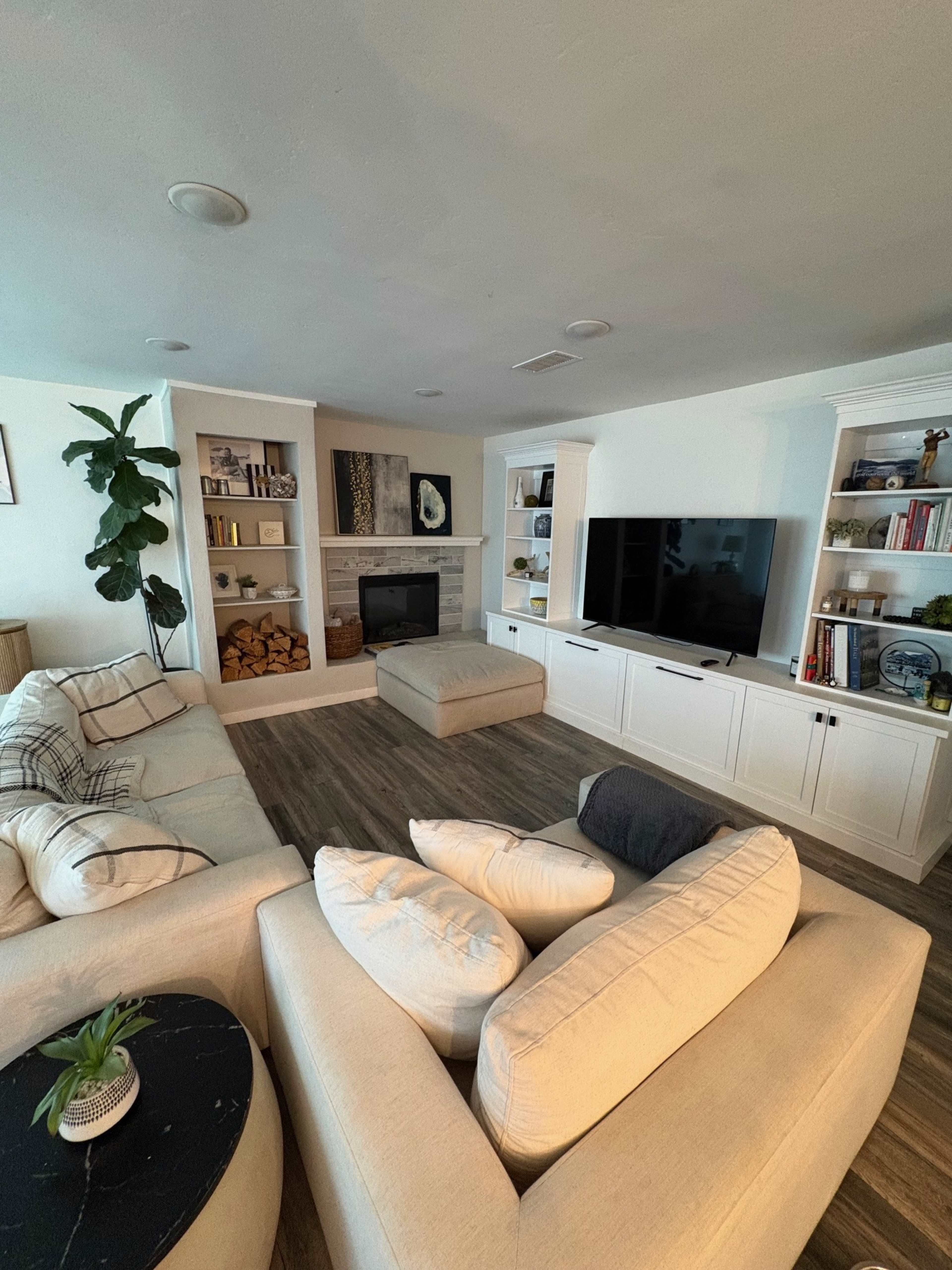 The living room features a sofa arrangement around a central ottoman, with built-in shelves, a fireplace, and a wall-mounted television.