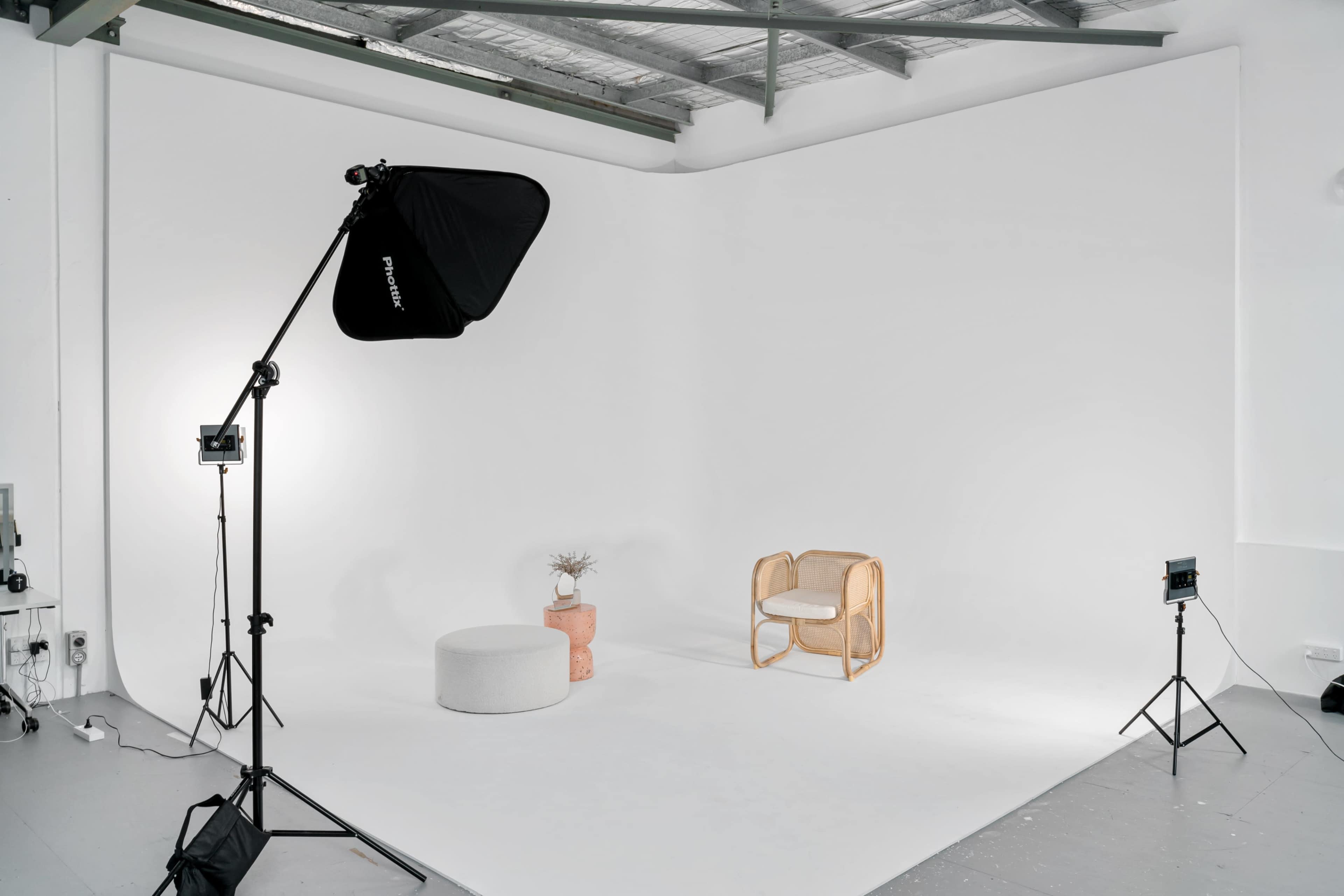 A photography studio features a simple setup with a chair, a round stool, and a potted plant against a white backdrop.