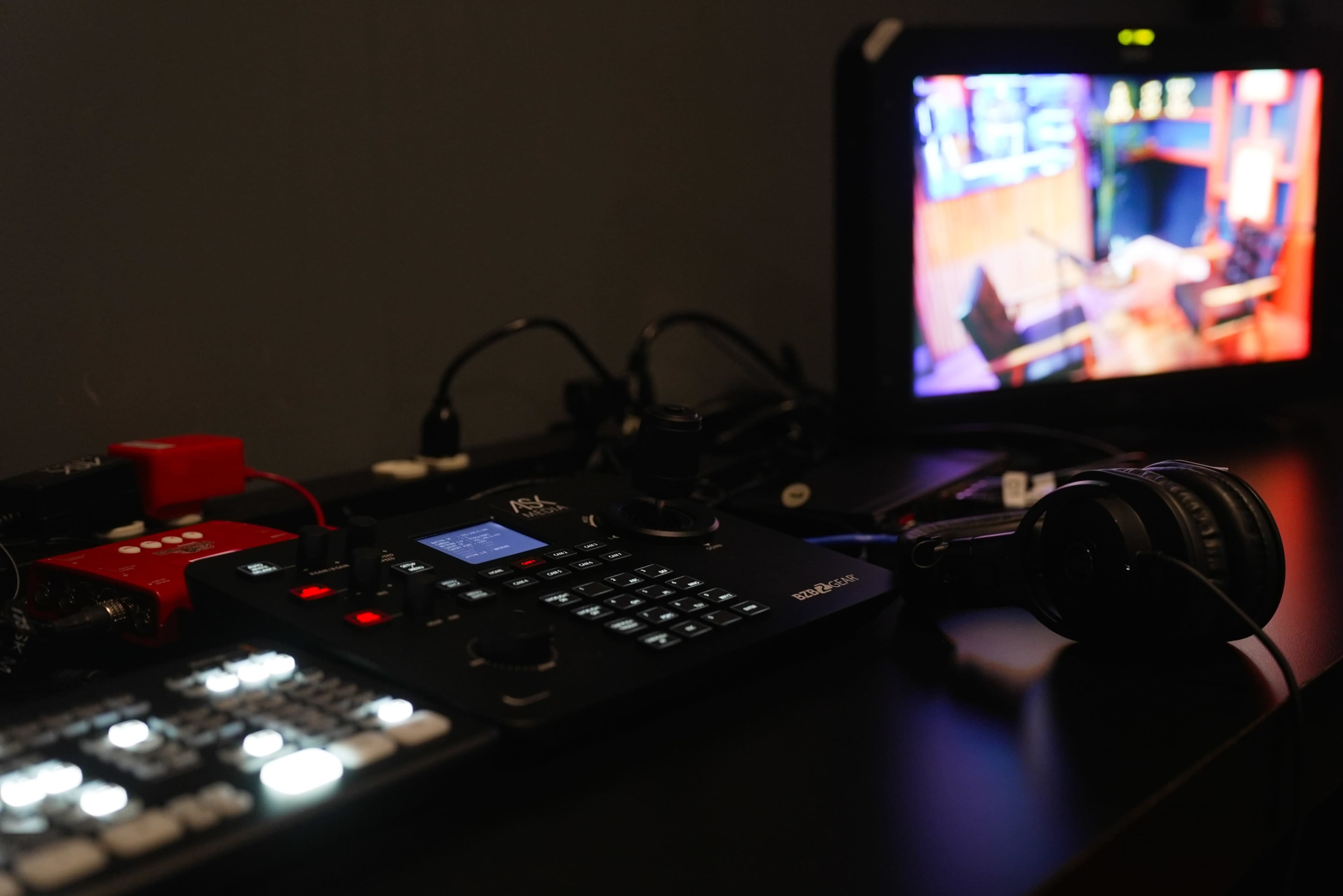 The image shows a control panel with various buttons and knobs, alongside a monitor displaying a well-lit studio setup and headphones resting on the table.